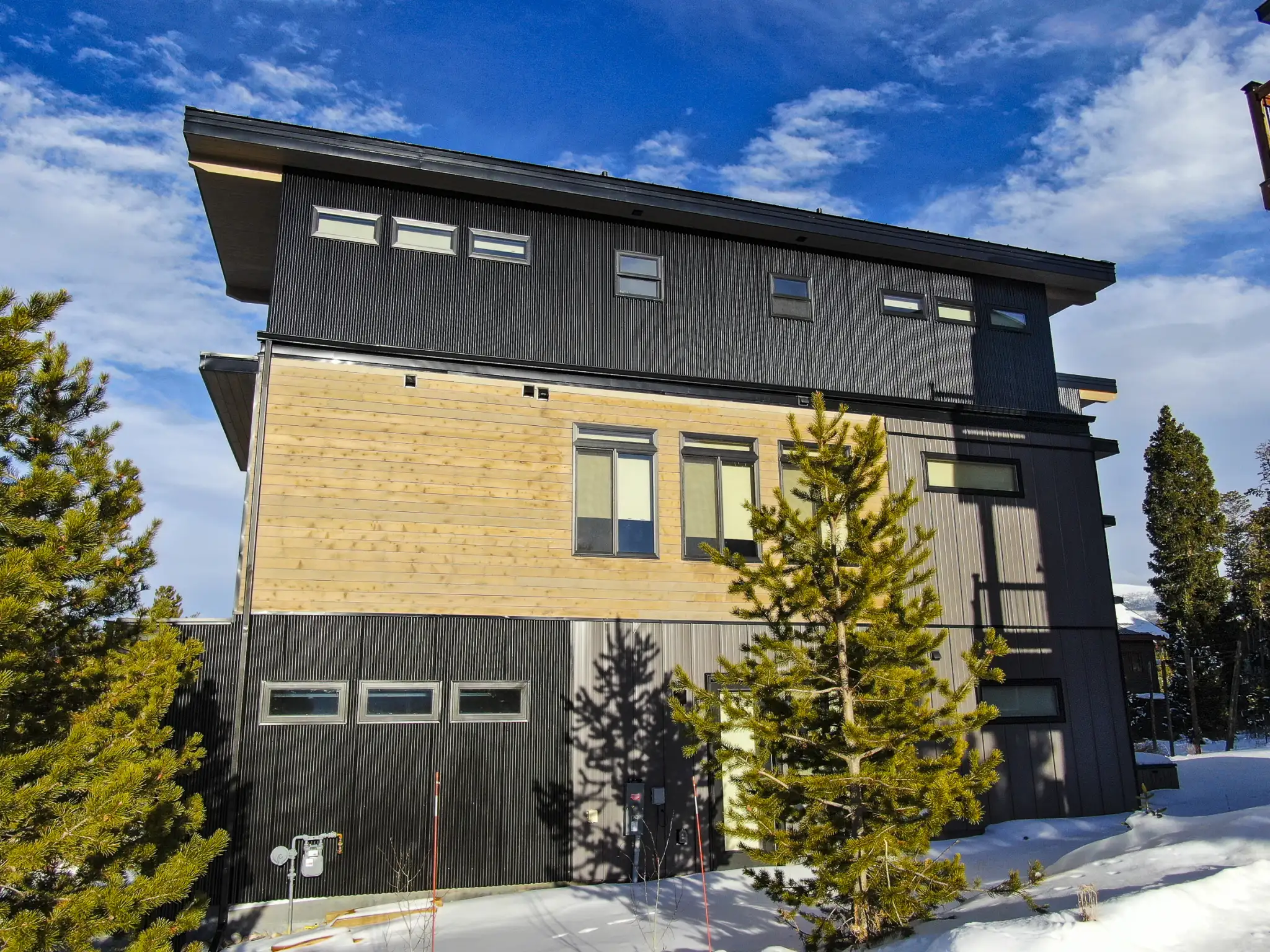 Three-story home under construction with dark upper siding, raw cedar middle section, two-car garage, snowy site with pine trees and blue sky