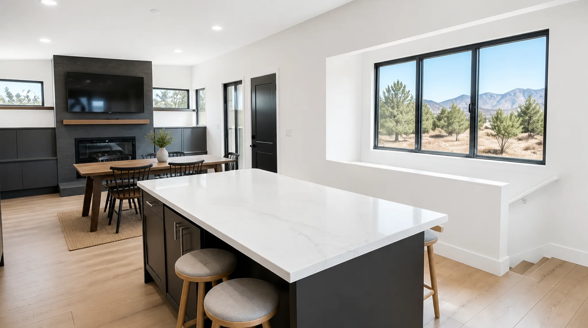 Open-plan kitchen and living area with dark charcoal island, white quartz countertop, round bar stools, black fireplace surround with TV, dining table with black spindle chairs, light wood floors, and black-framed windows with pine and mountain views
