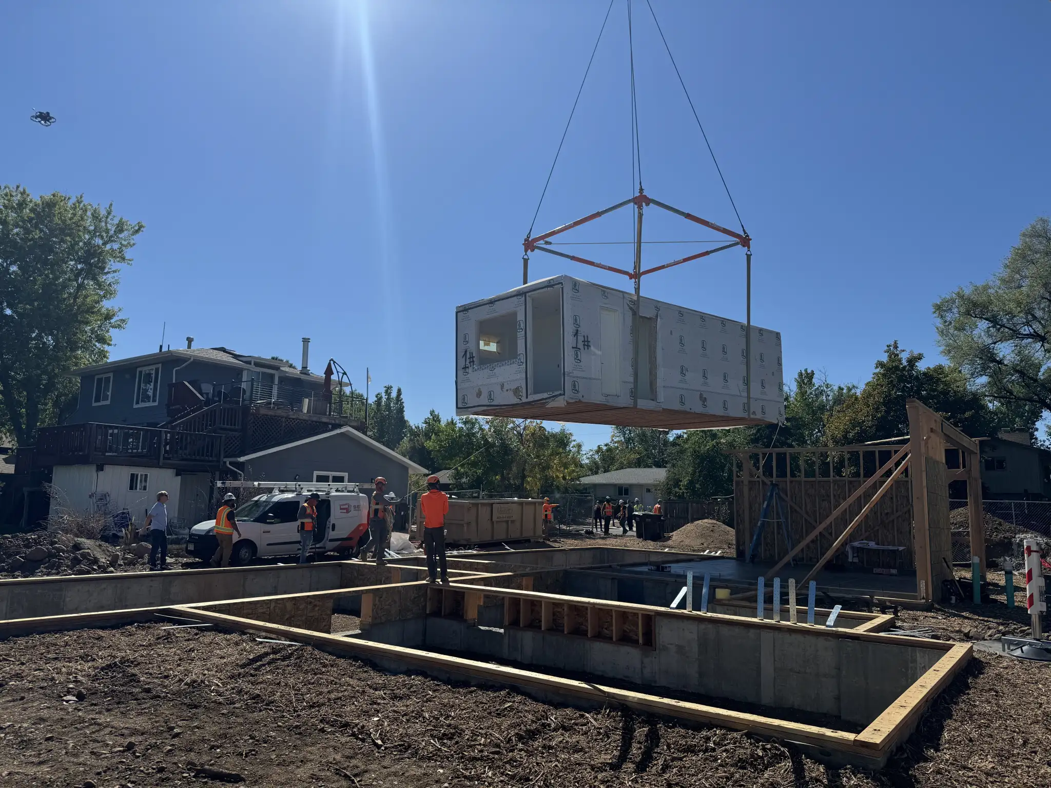 Construction crew in safety vests using a crane to lower a modular home unit onto a concrete foundation in a residential neighborhood