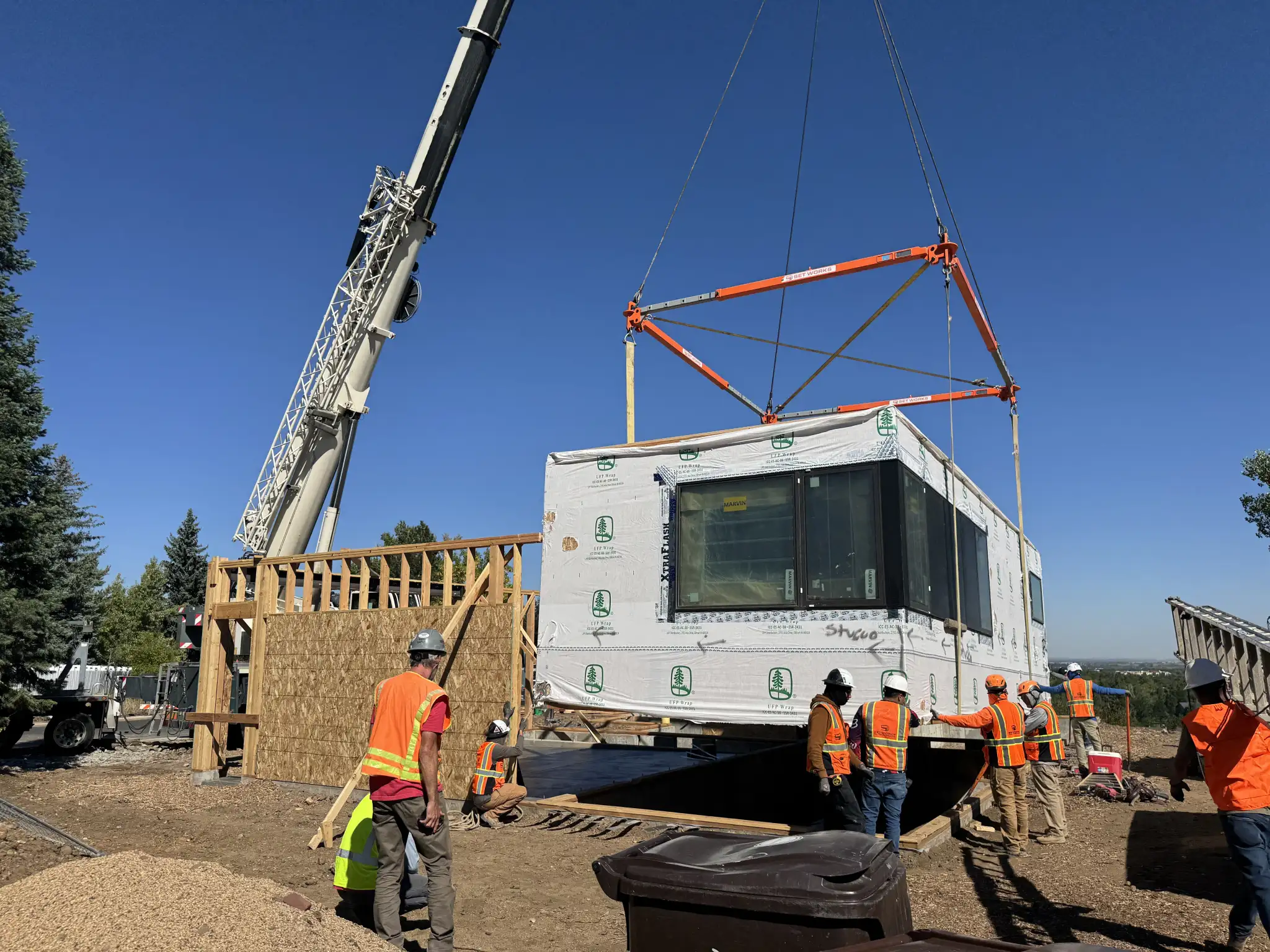 Large crane lowering a modular home module as crew members in orange safety vests guide the placement on site