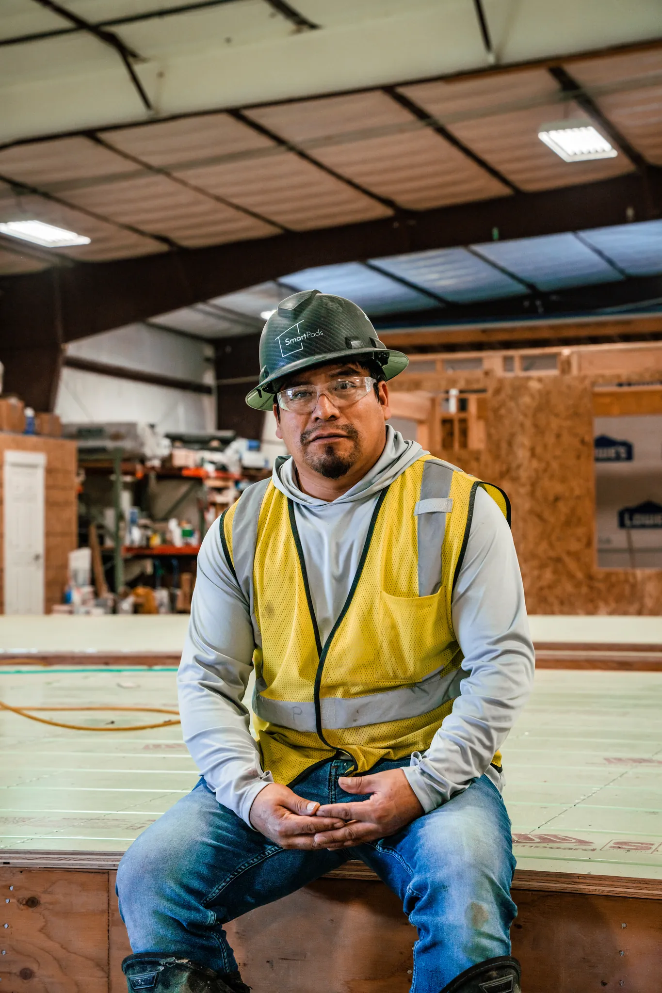 Smartpads worker in hard hat and safety vest seated on a structural panel inside the modular home factory