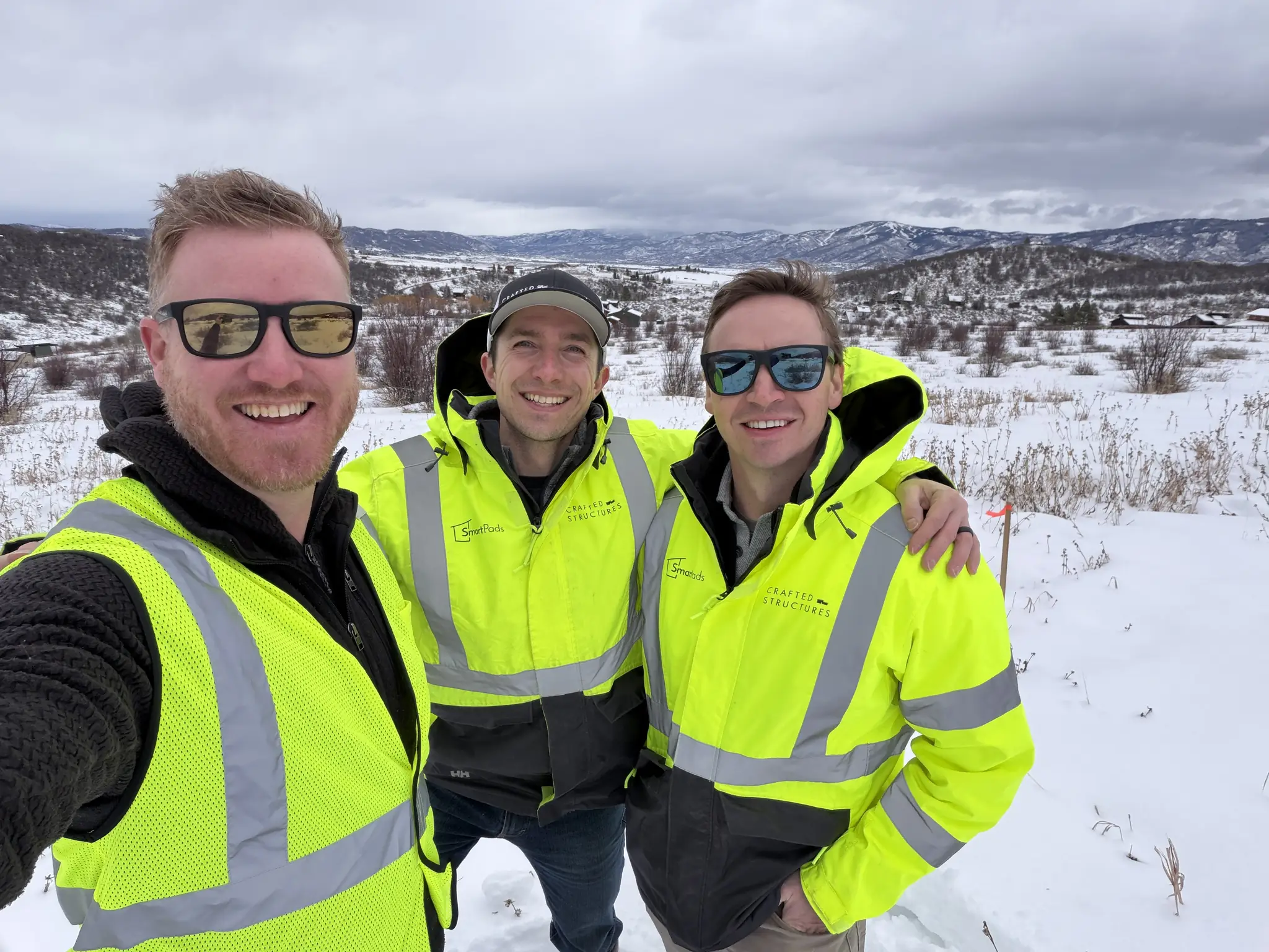 Three Smartpads team members in yellow safety vests taking a selfie on a snowy job site with mountain valley views