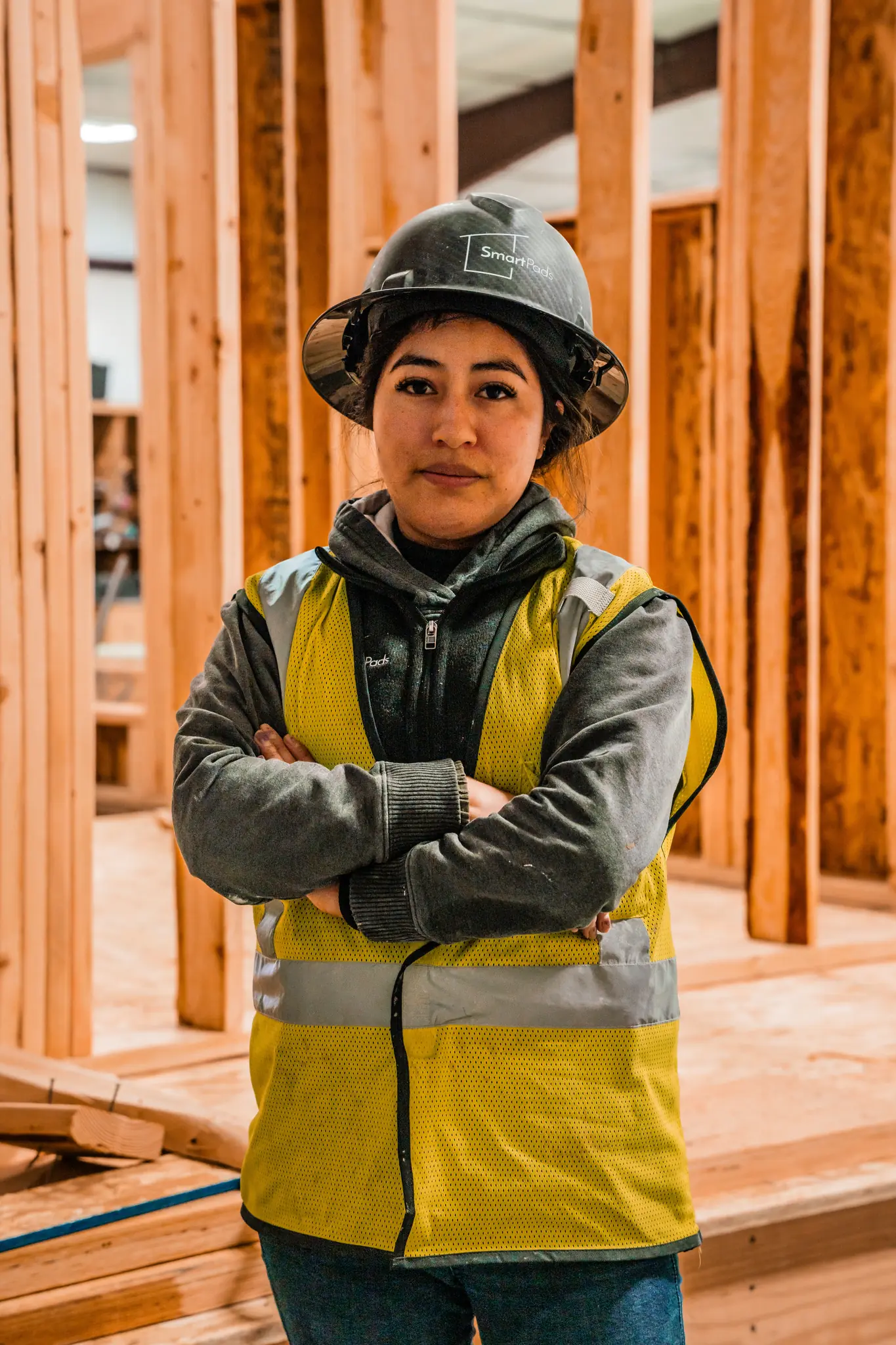Smartpads worker in hard hat and yellow safety vest standing among wood wall framing panels in the factory