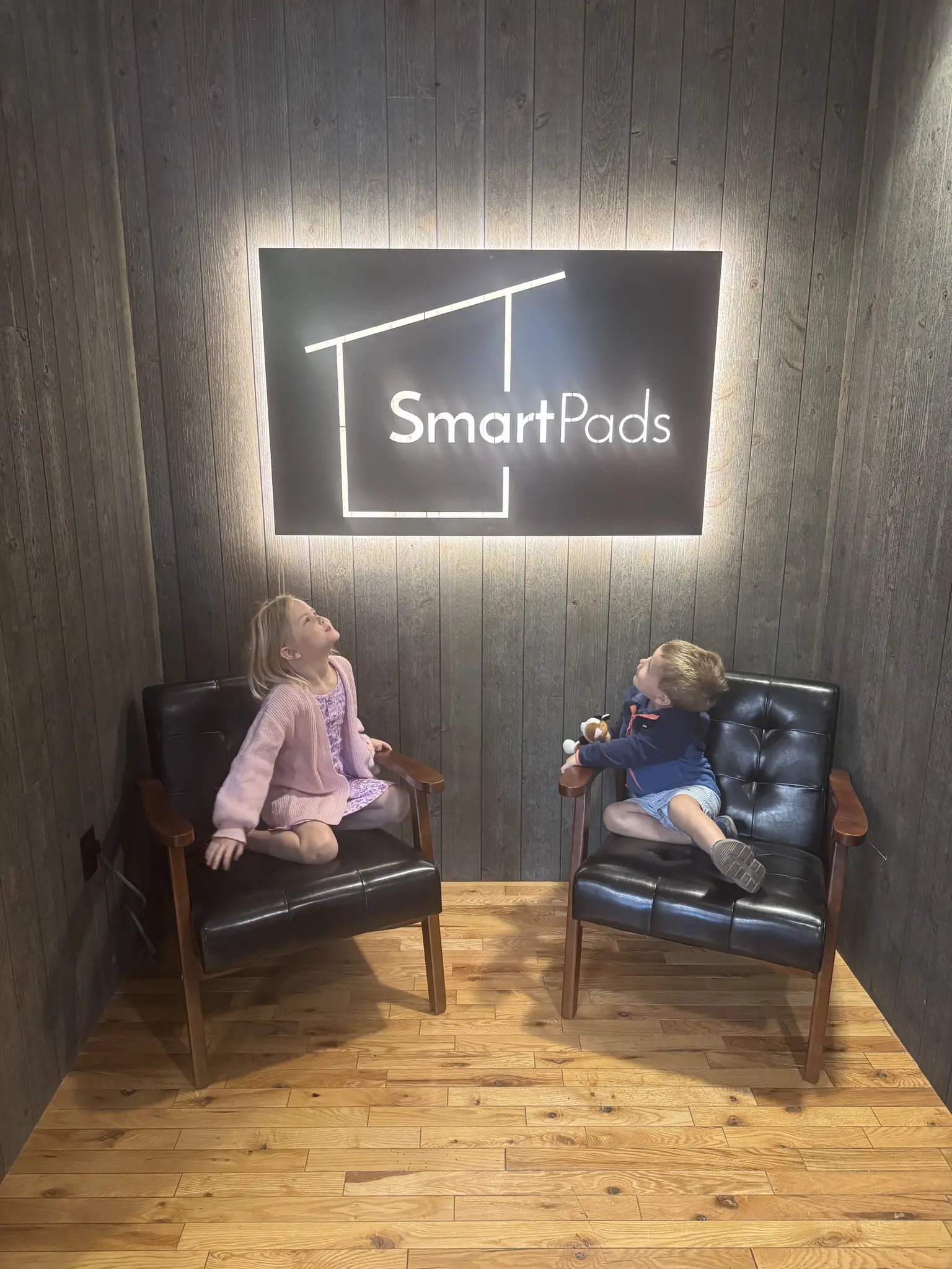 Two young children sitting in leather chairs beneath an illuminated Smartpads logo sign in the company showroom
