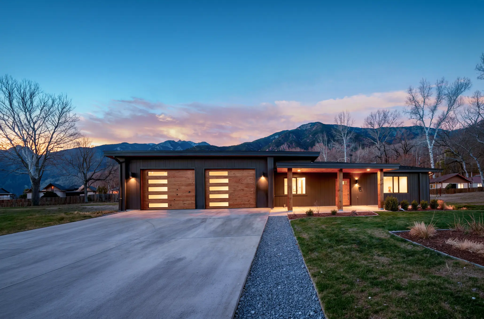 Single-story ranch-style home with dark exterior, cedar-paneled garage doors, and wide driveway set against foothills at sunset