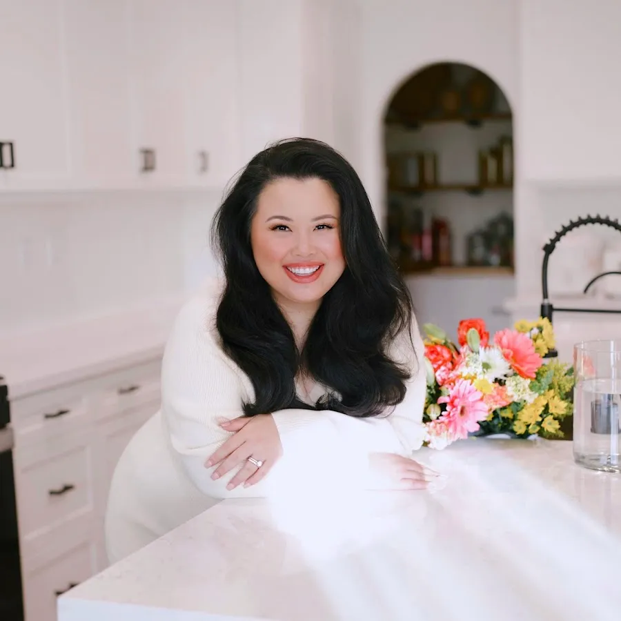 Smiling woman with long dark hair leaning on a white kitchen counter with a colorful bouquet of flowers nearby.
