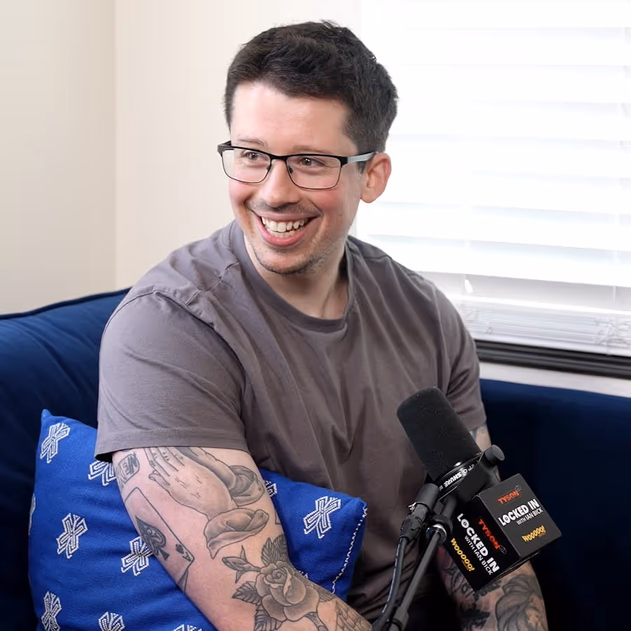 Smiling man with glasses and tattoos on his arms sitting on a blue couch with a microphone labeled 'LOCKED IN'.