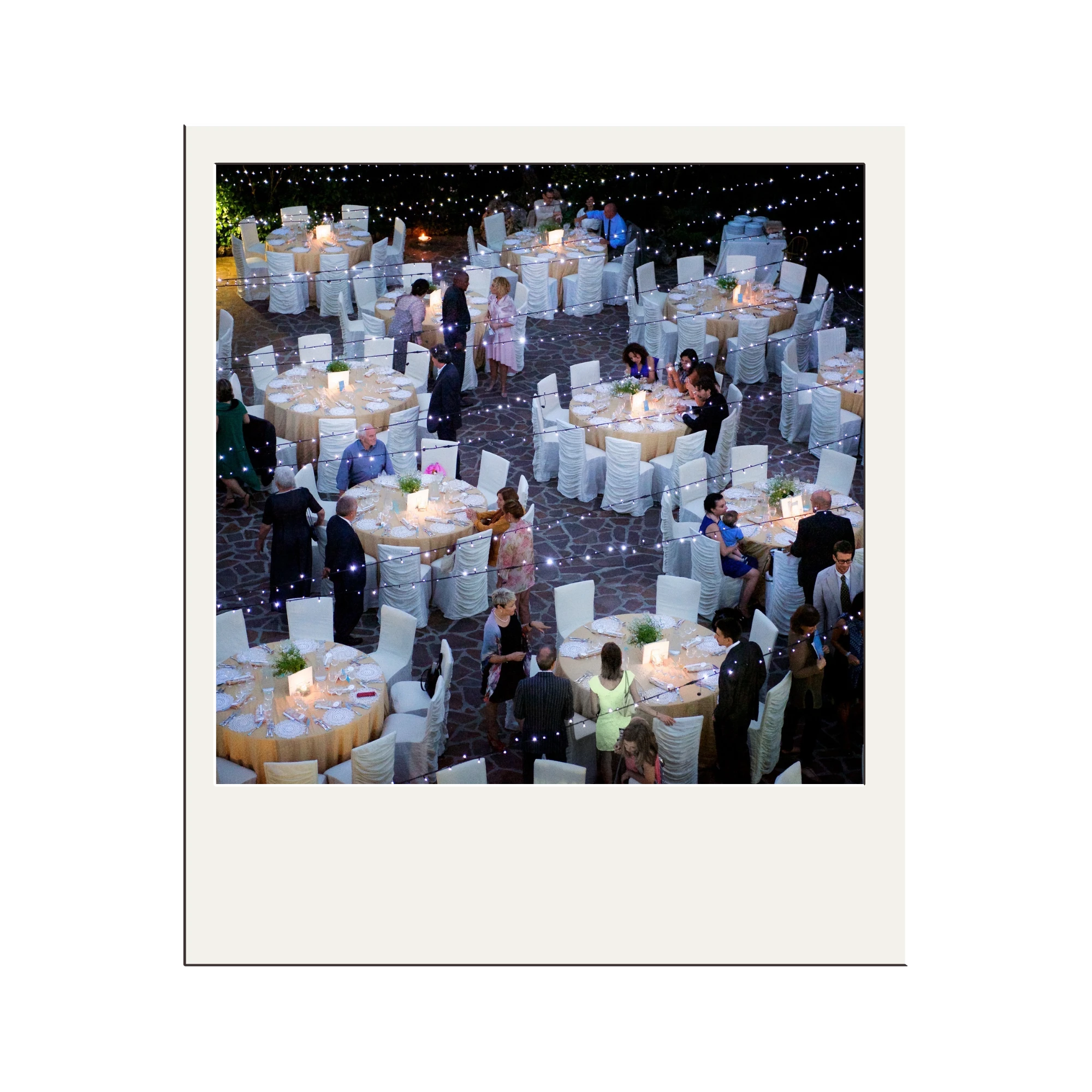 Overhead view of an outdoor evening event with round tables covered in beige tablecloths, white chairs, string lights overhead, and guests mingling.