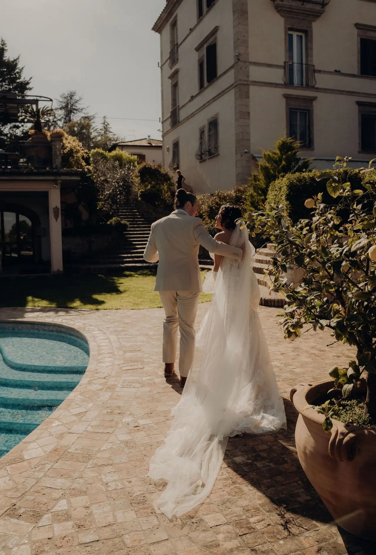 Bride in a white dress and veil walking arm in arm with groom in a light-colored suit on a stone pathway beside a swimming pool and greenery.