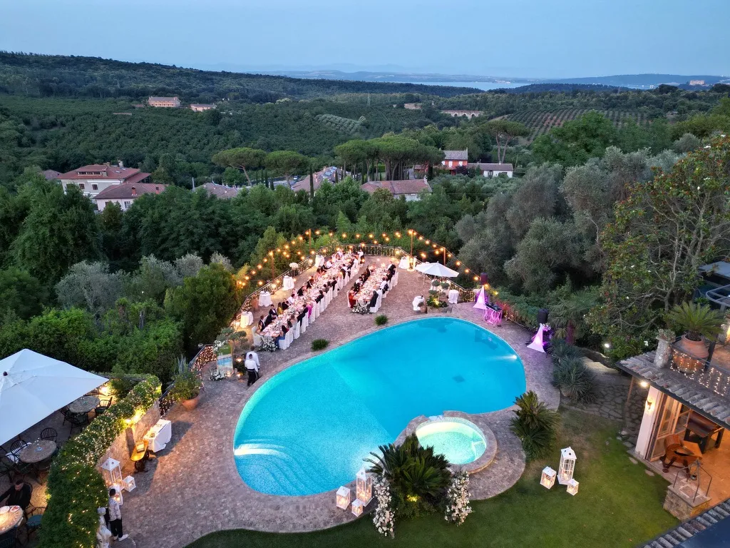Evening outdoor event with long decorated tables and string lights beside a kidney-shaped swimming pool and hot tub, surrounded by lush greenery and distant hills.