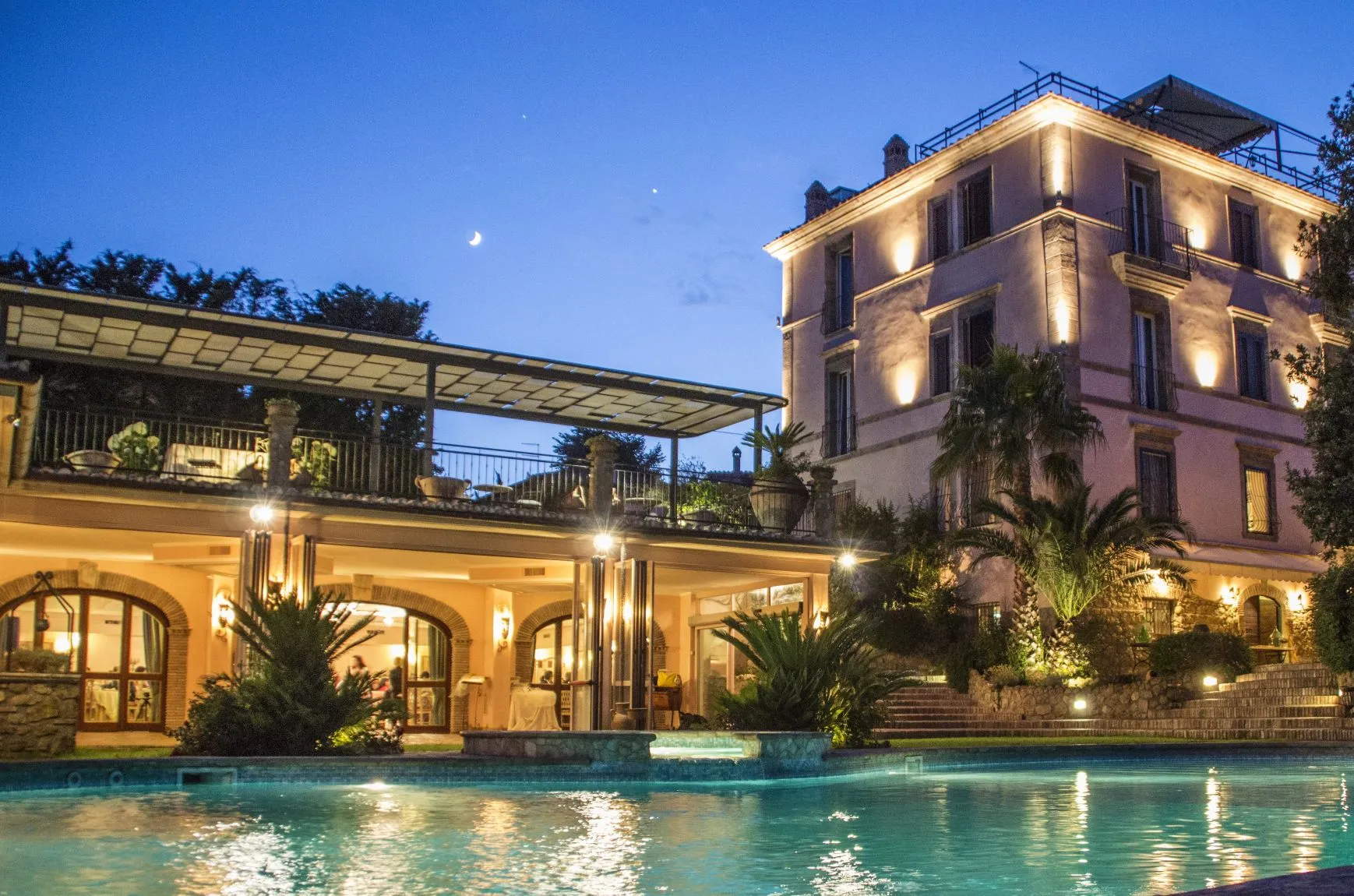 Elegant illuminated villa with a rooftop terrace and a swimming pool at dusk under a crescent moon.