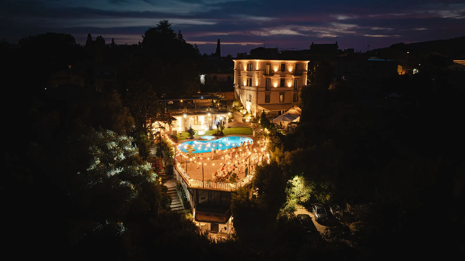 Night view of an illuminated villa with a swimming pool and outdoor dining area decorated with string lights.