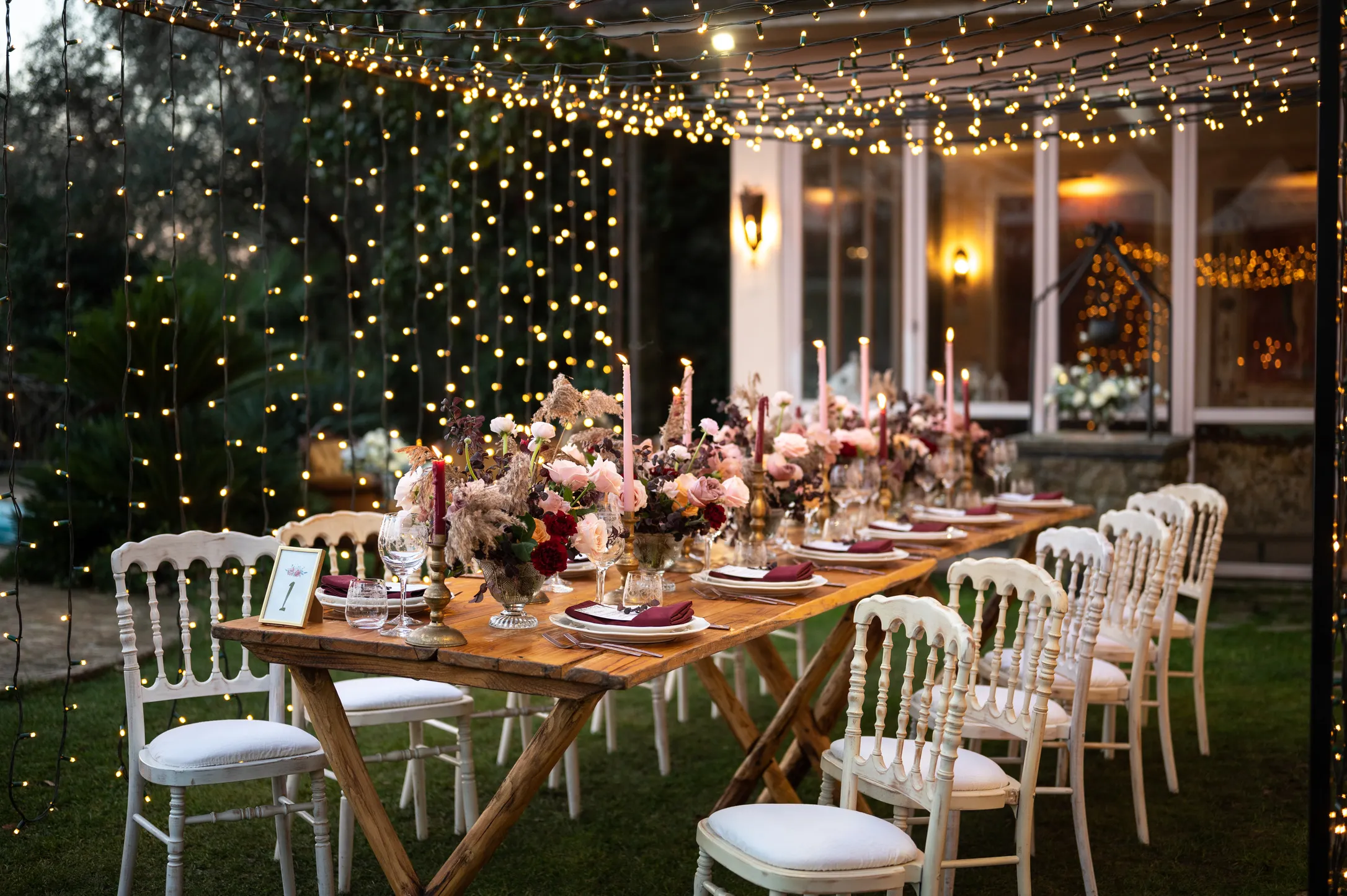 Elegant outdoor dining table set with floral centerpieces, candles, and string lights overhead in a garden setting at dusk.