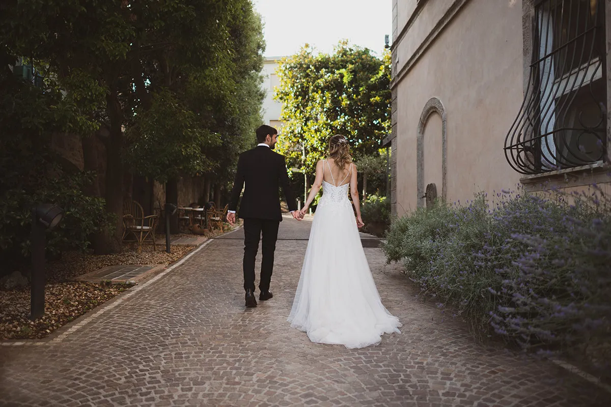 Bride in a white wedding dress and groom in a black suit holding hands walking down a cobblestone path lined with trees and a building.