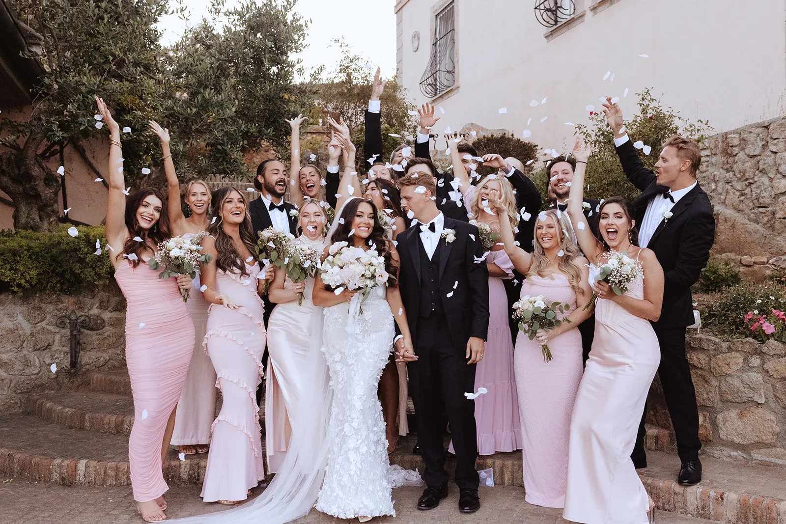 Bride and groom holding hands surrounded by bridesmaids in pink dresses and groomsmen in black suits tossing white flower petals.