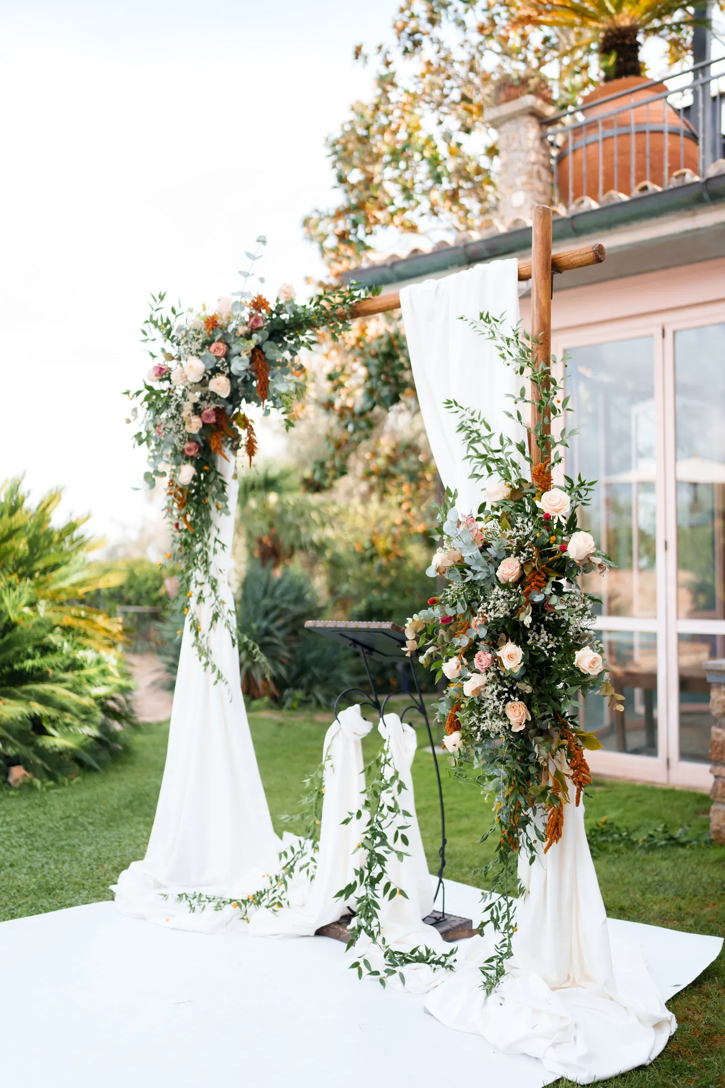 Outdoor wedding altar with a wooden arch draped in white fabric and decorated with lush greenery and pastel roses.