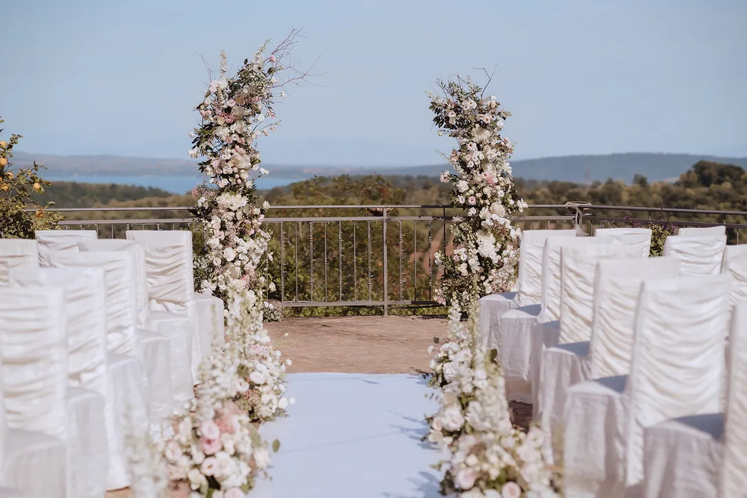 Outdoor wedding ceremony setup with white chairs and floral arches overlooking a scenic landscape with hills and water in the background.