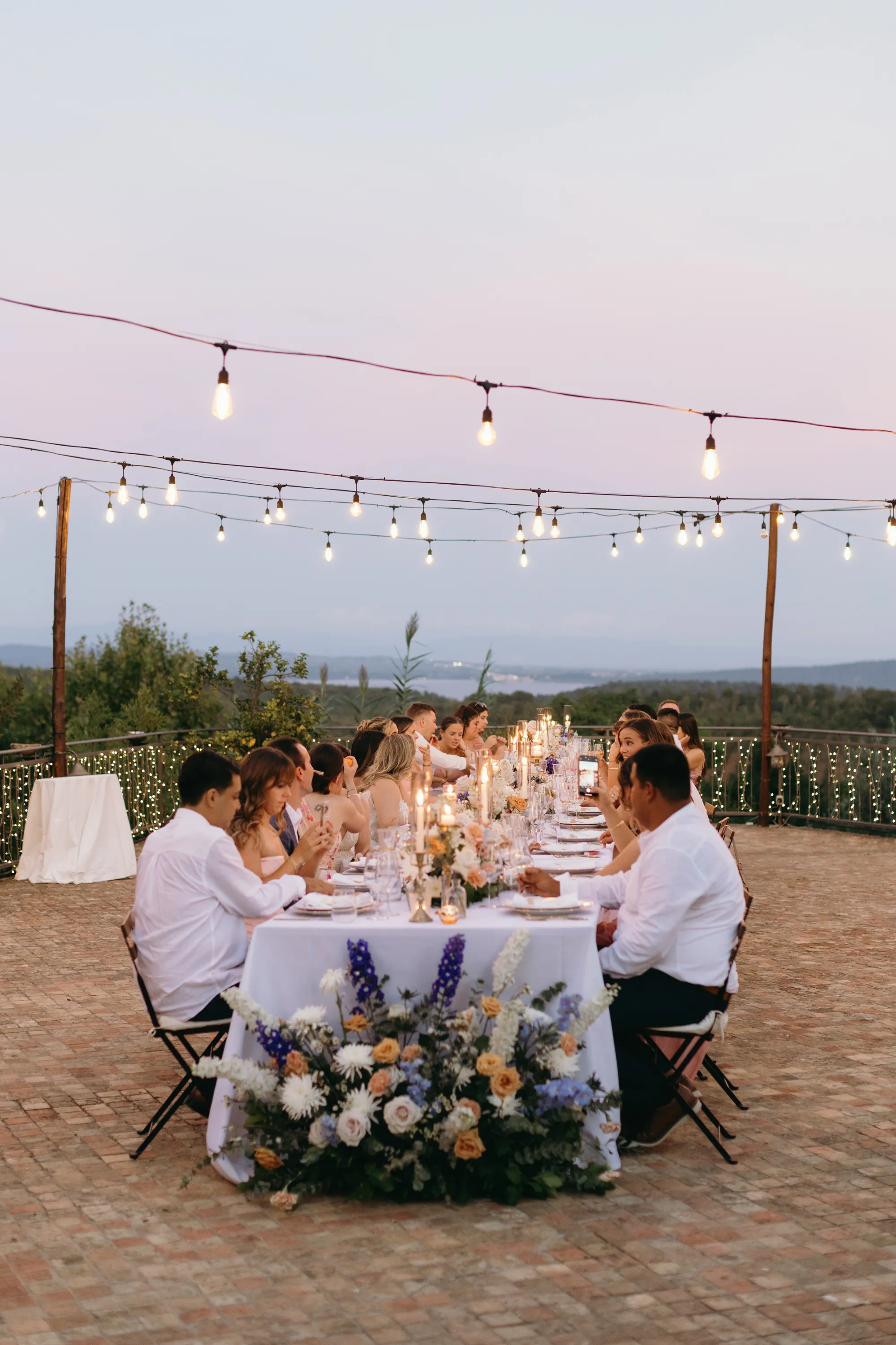 Outdoor dinner party with guests seated at a long table adorned with candles, flowers, and string lights overhead during dusk.