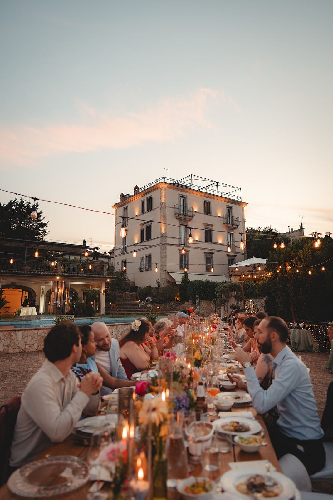 Long outdoor dining table with guests enjoying a sunset dinner party near a lit villa and string lights.