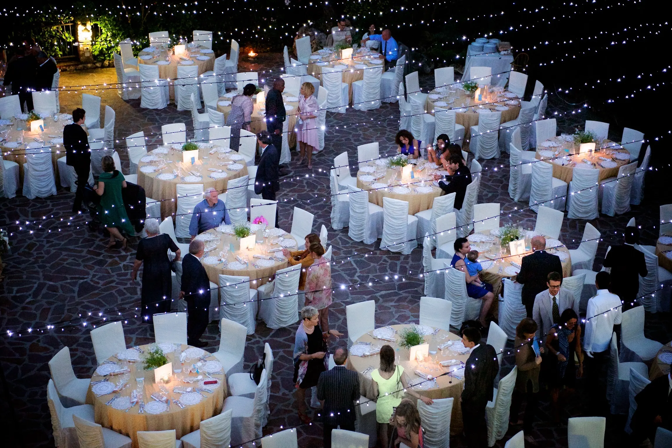 Outdoor evening event with round tables covered in beige tablecloths, white chairs, and string lights overhead.