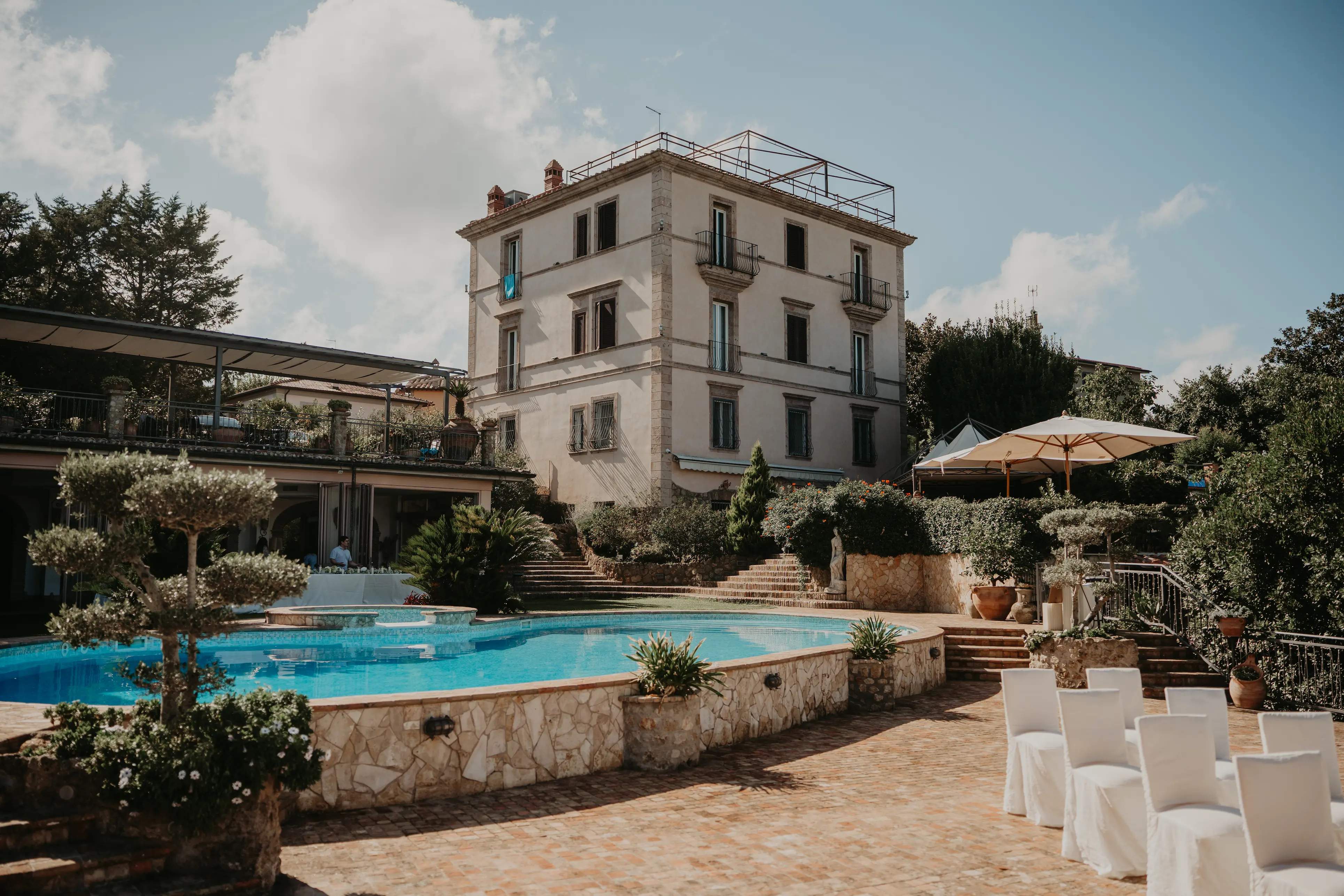 Elegant stone villa with a blue swimming pool, white chairs, and umbrellas in a sunny garden setting.