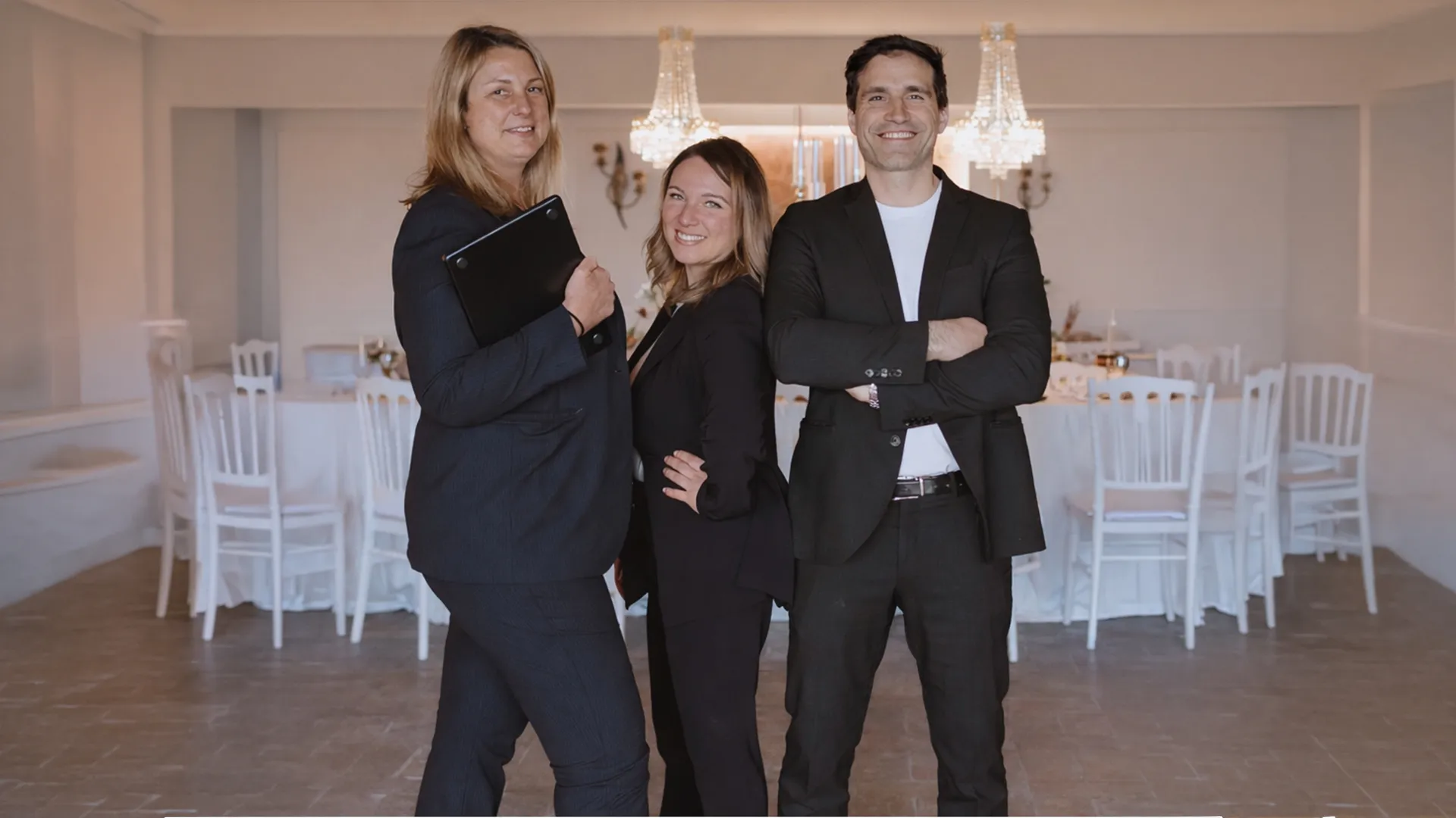 Three professionals in business attire posing confidently in front of a dining table with crystal chandeliers in the background.