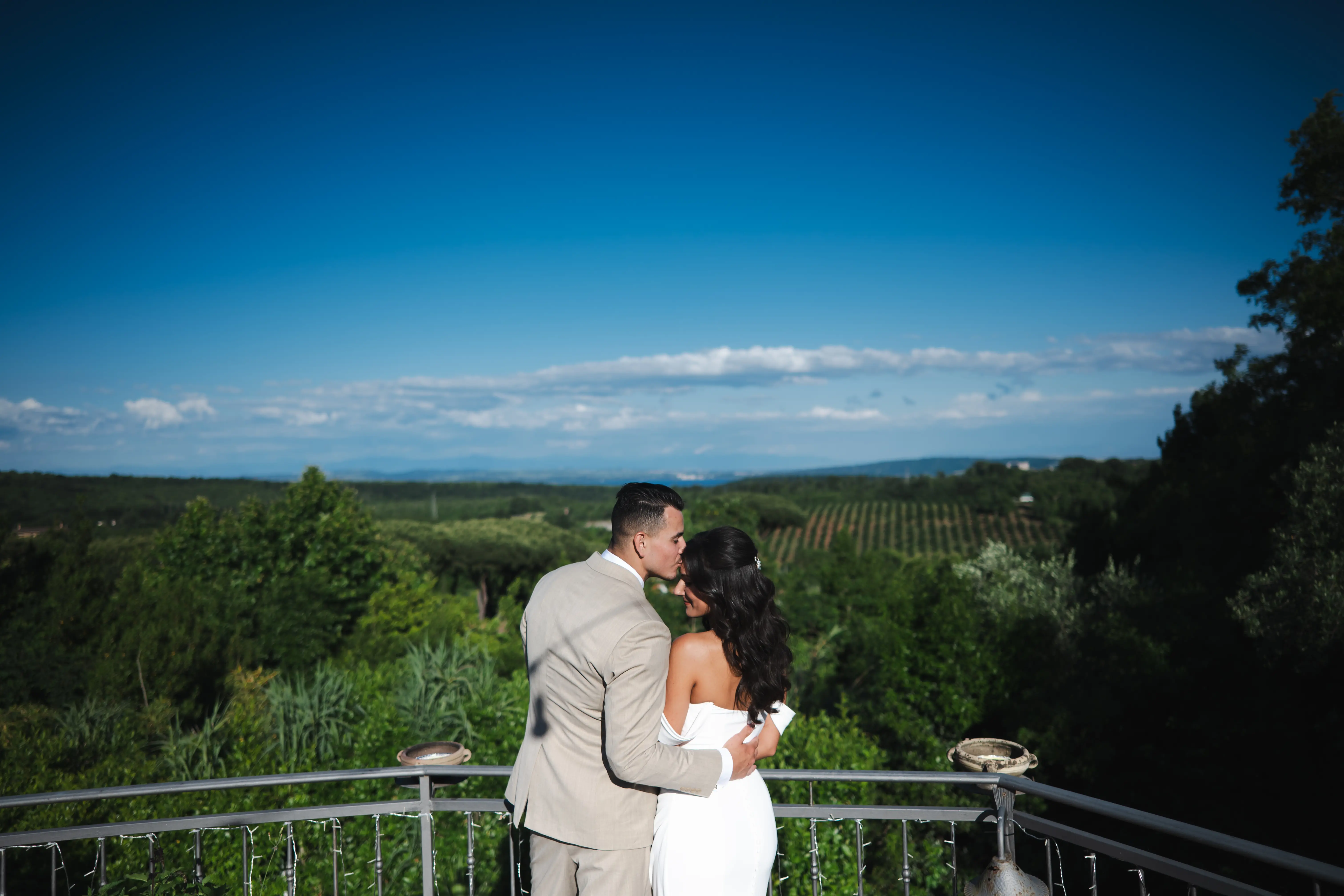 Bride and groom embracing on a balcony overlooking green fields and a clear blue sky.