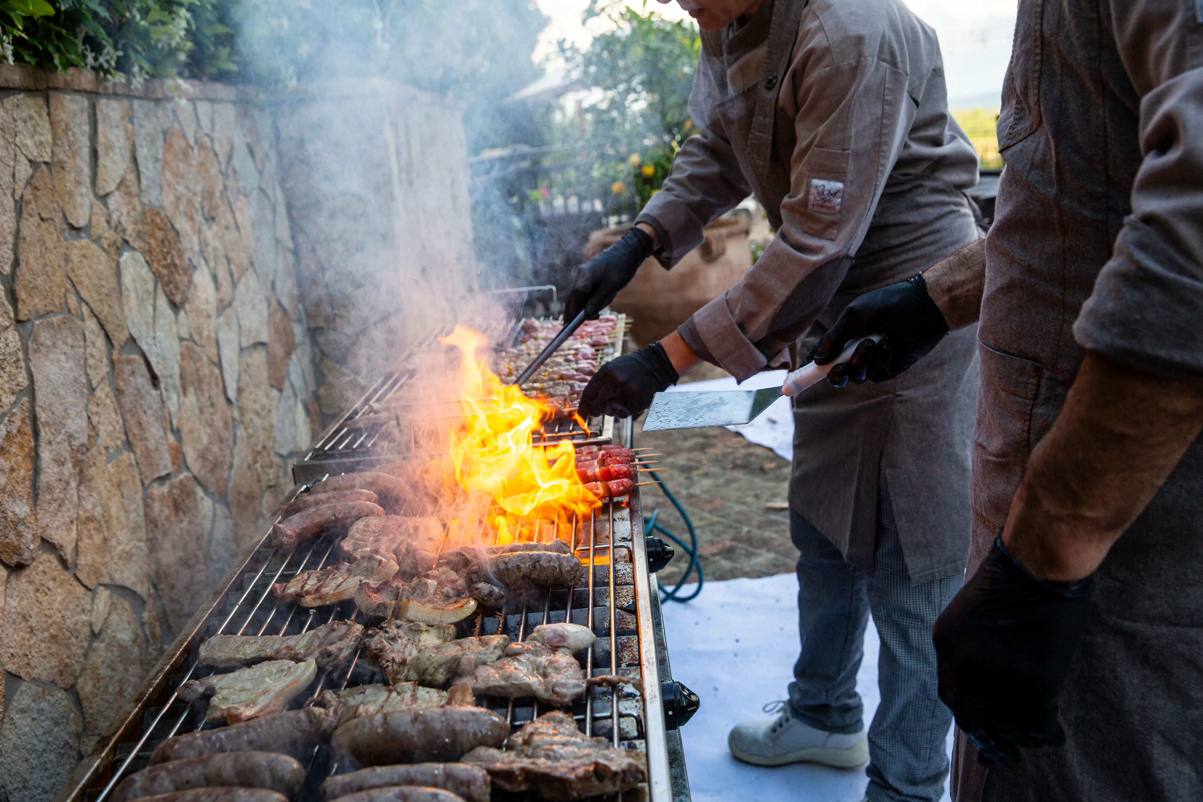 Two chefs grilling various meats and skewers over an outdoor flame grill with smoke rising.