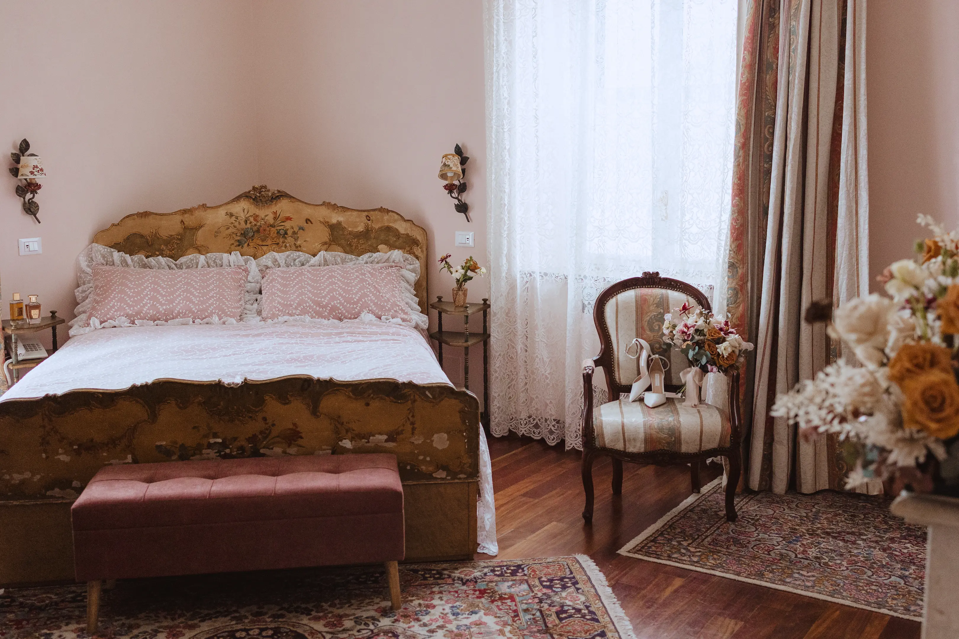 Vintage bedroom with an ornate floral headboard bed, pink pillows, lace curtains, an upholstered chair holding bridal shoes and bouquet, and floral rugs on wooden floor.