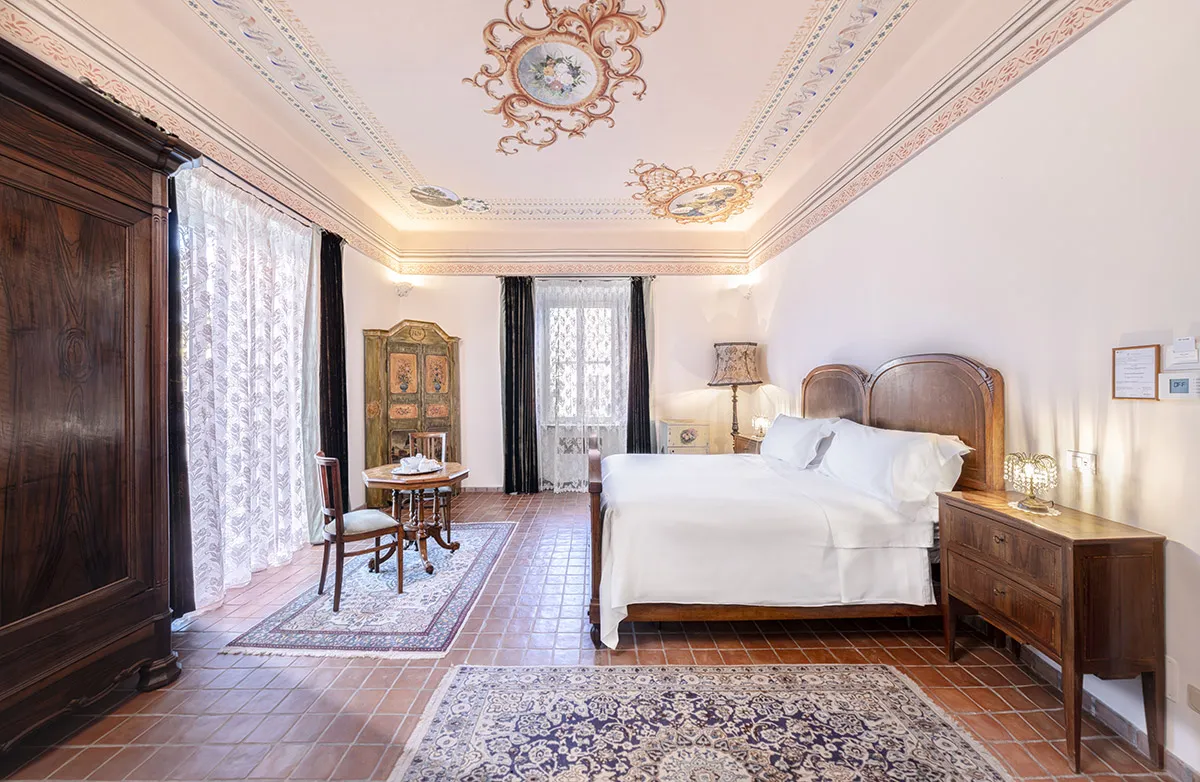 Bright vintage bedroom with ornate ceiling, wooden bed with white linens, antique wooden wardrobe, small table with two chairs, and decorative rugs on tiled floor.