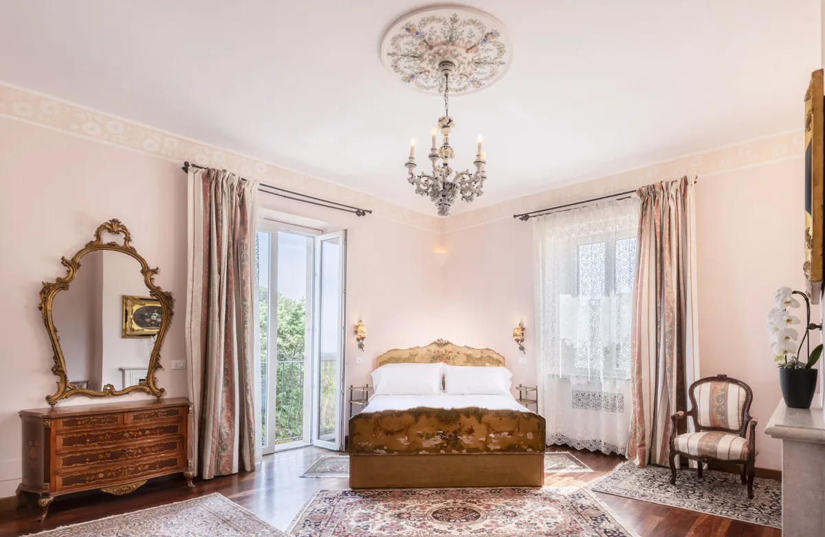 Bright bedroom with vintage-style bed, ornate ceiling chandelier, wooden dresser with gold-framed mirror, and floral curtains.