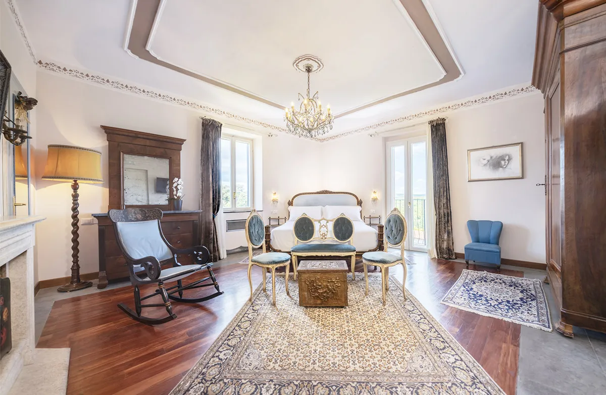 Spacious elegant bedroom with a large bed, antique chairs, a wooden rocking chair, classic rug, chandelier, and tall windows with curtains.