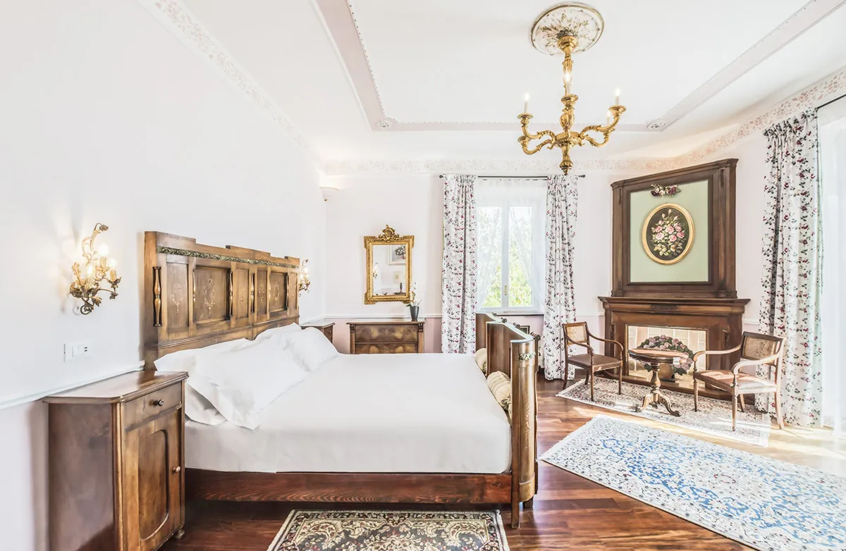 Bright, spacious bedroom with a large wooden bed, floral curtains, vintage wooden furniture, and a decorative fireplace with floral artwork.