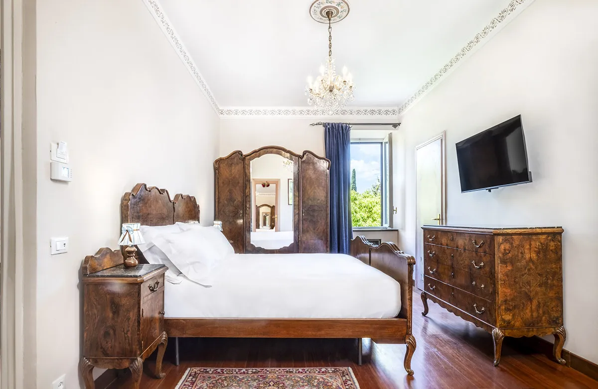 Classic bedroom with wooden bed, nightstand, dresser, wall-mounted TV, and open window with blue curtain.