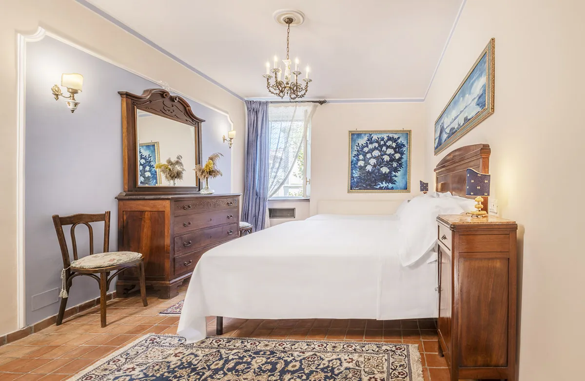 Bright bedroom with white bed linens, wooden headboard and furniture, a large mirror dresser, floral paintings, and a chandelier.