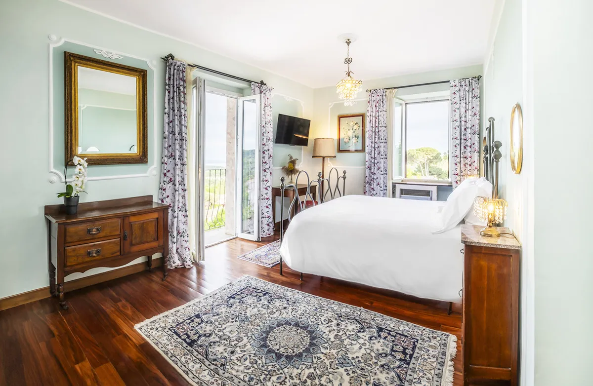 Bright bedroom with a white bed, floral curtains, wooden furniture, two windows, and a balcony door.