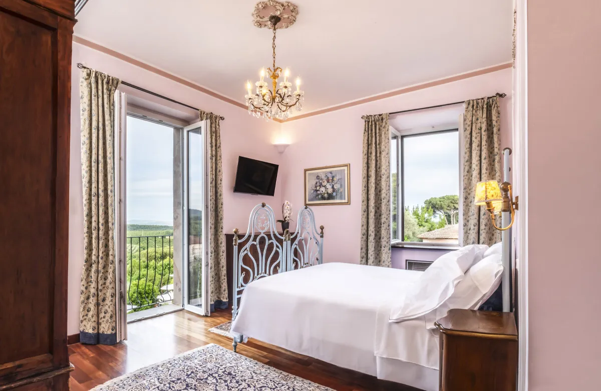 Bright bedroom with a double bed with white linens, two windows with floral curtains, wooden furniture, a chandelier, and a flat-screen TV.