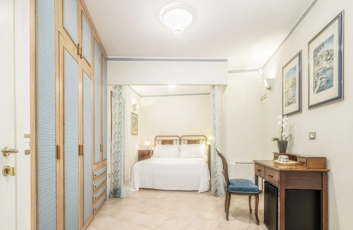 Bright bedroom with a white bed, wooden wardrobe with blue panels, vintage wooden desk and chair, and framed coastal artwork on the wall.