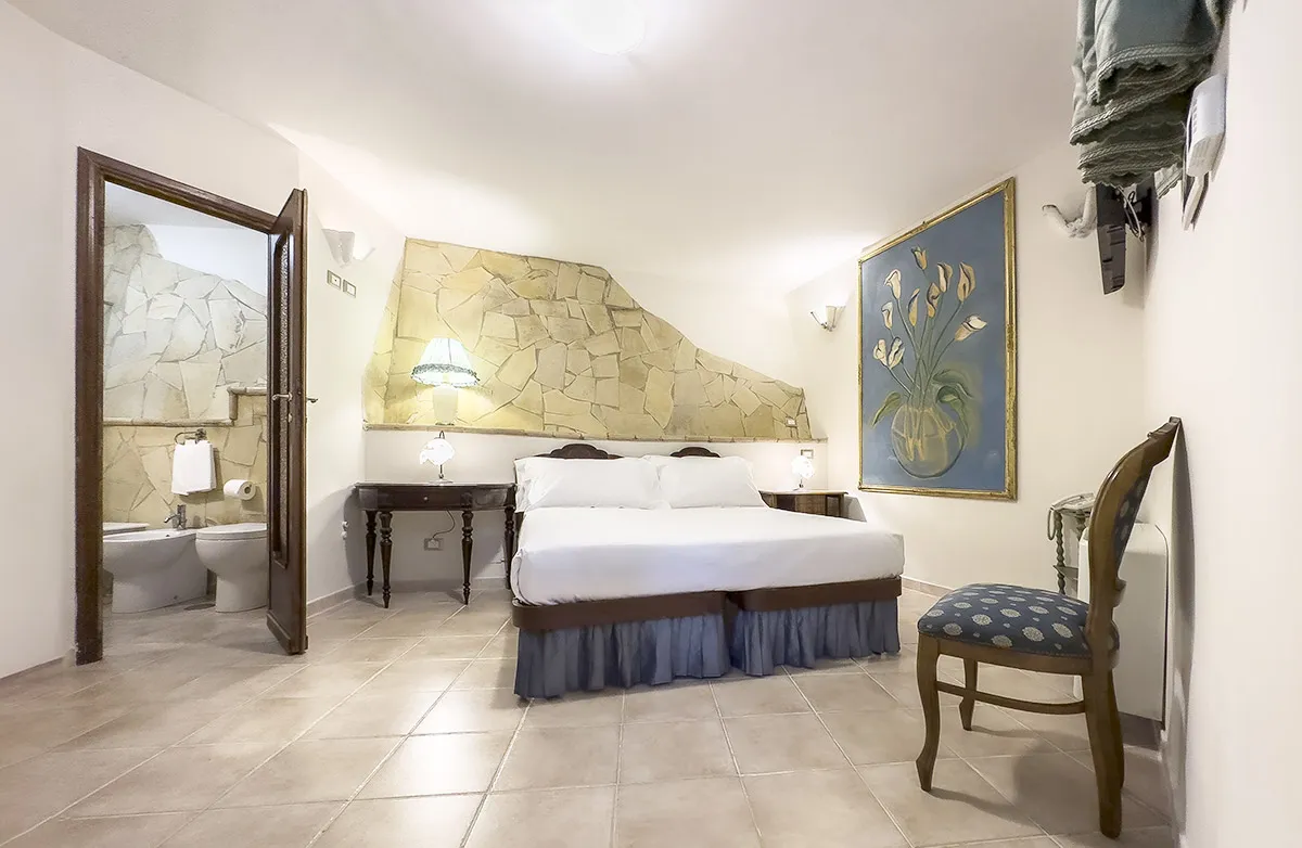Bright hotel room with a large bed, two bedside tables with lamps, stone accent wall, floral painting, wooden chair, and open door showing tiled bathroom.