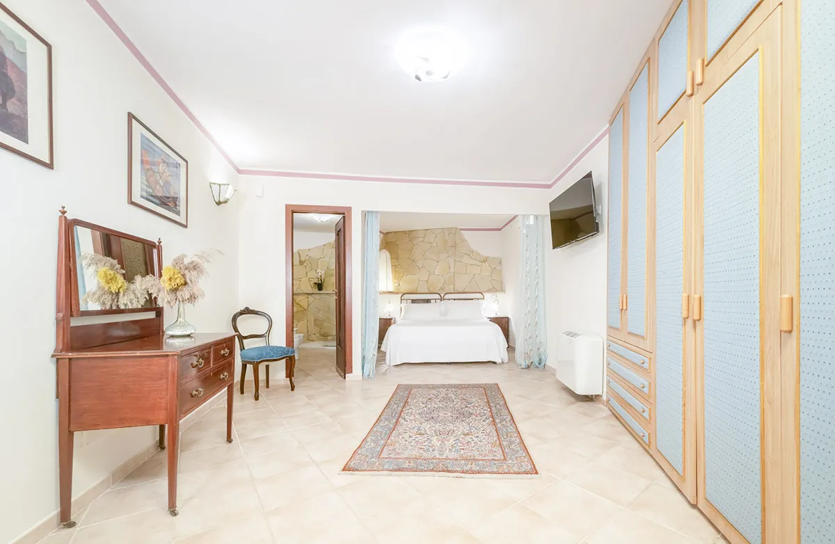 Bright hotel room with a double bed, stone accent wall, wooden wardrobe with blue panels, wooden dresser with mirror, vintage chair, and a wall-mounted TV.