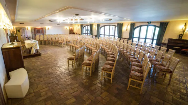 Large room with curved rows of wooden chairs arranged for a meeting or event, featuring wooden floors and windows with curtains.