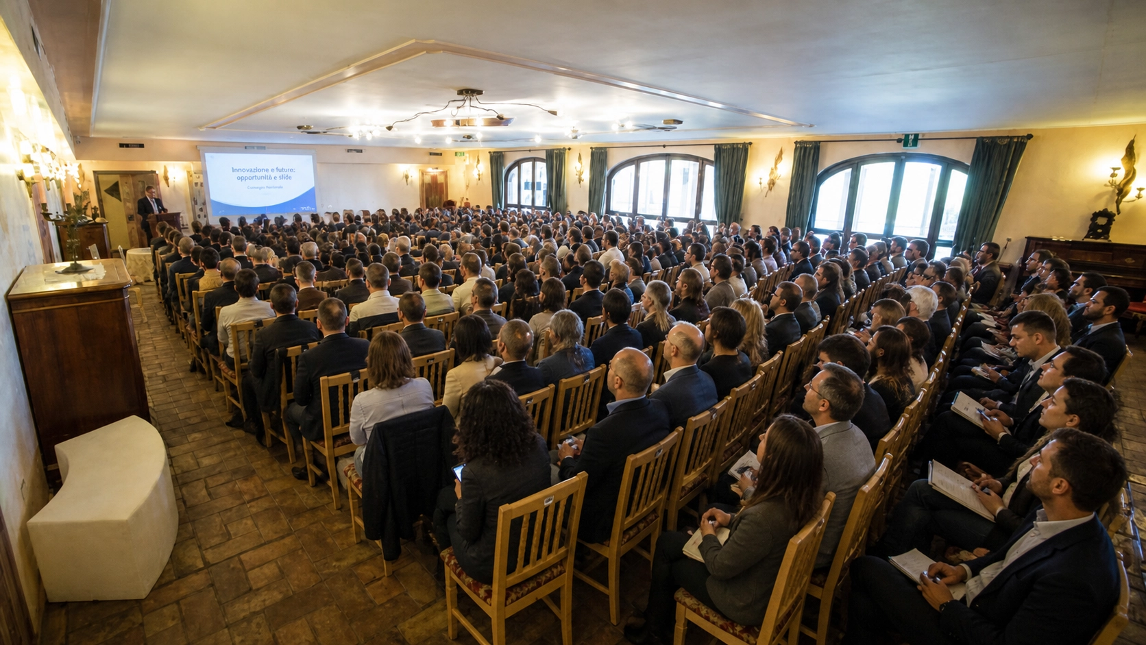 Large audience seated in rows listening to a speaker presenting a session titled 'Innovazione e future: opportunità e sfide' in a conference room with ornate windows and warm lighting.
