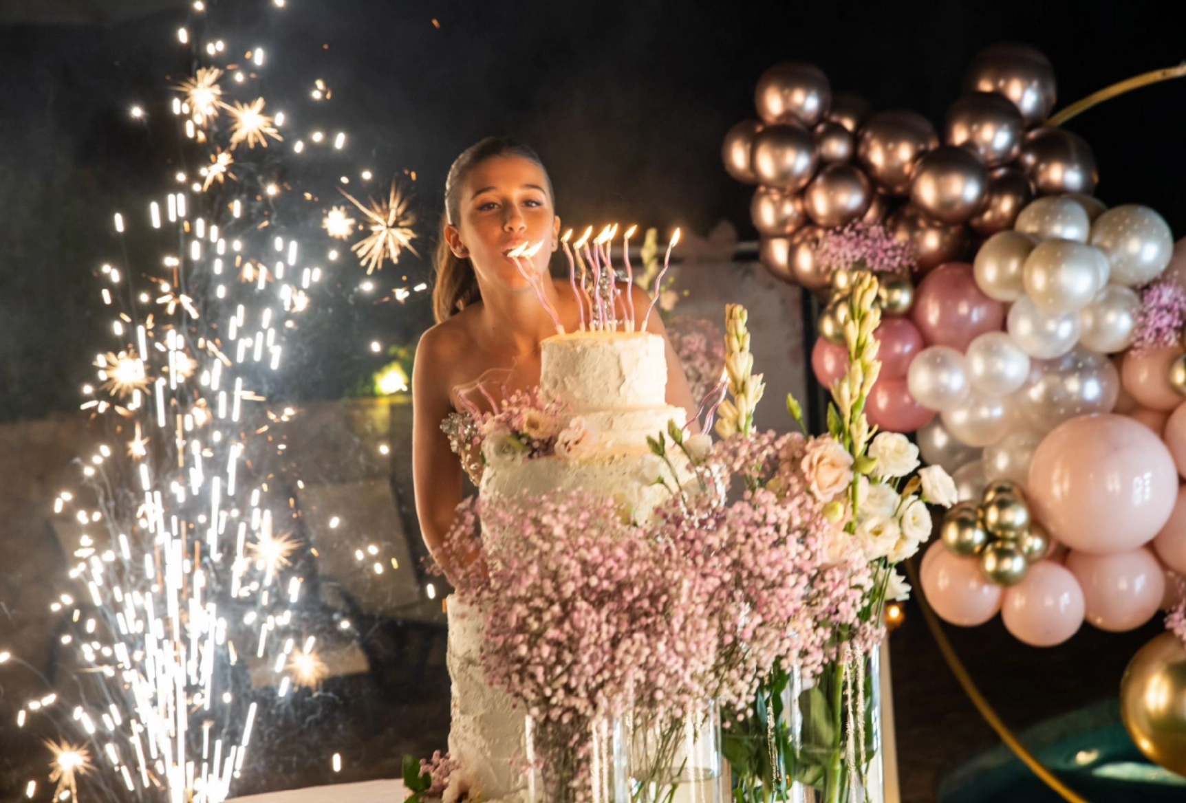 Girl blowing out candles on a tall white tiered birthday cake surrounded by pink flowers and lit sparklers, with pink and gold balloons in the background.