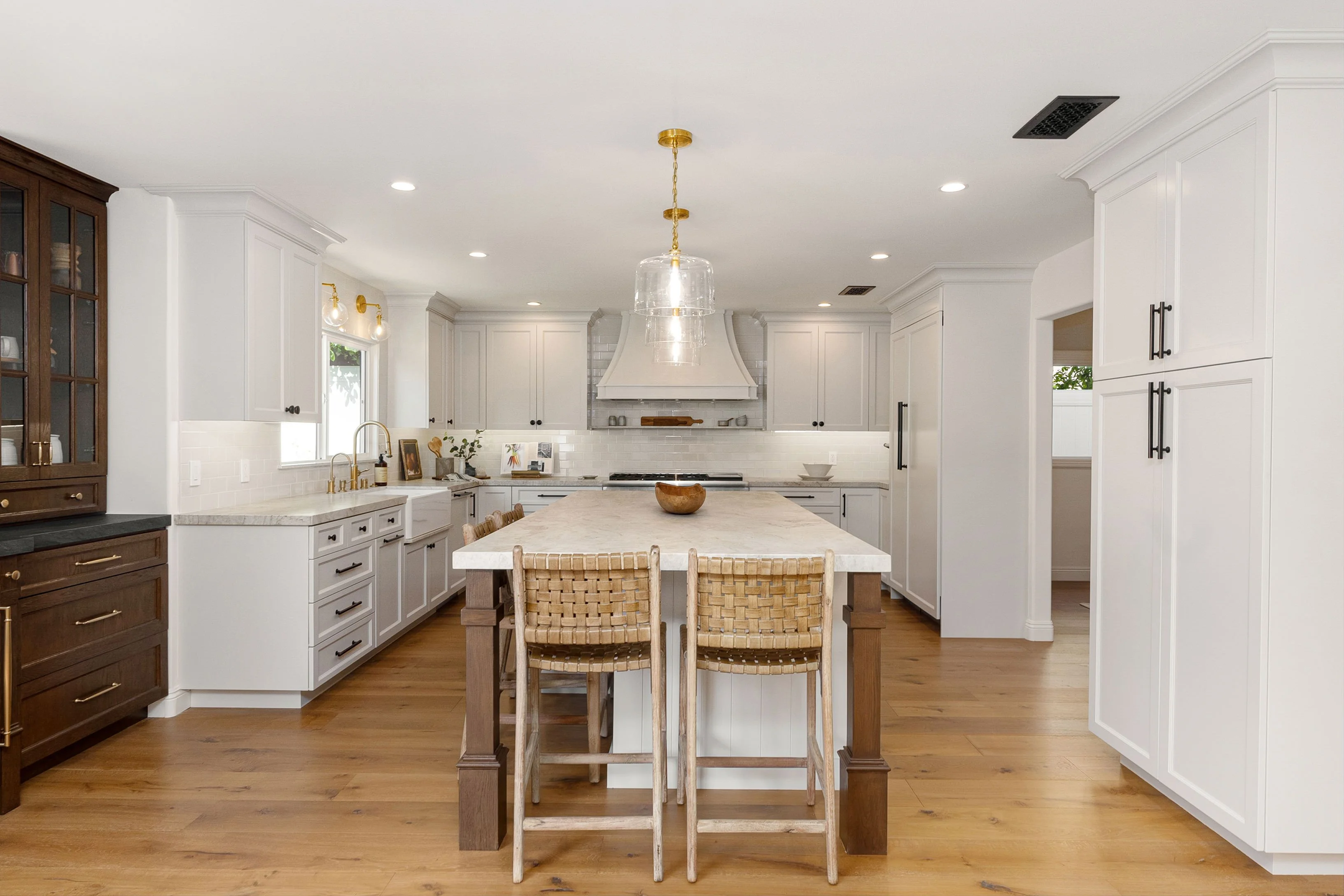 Modern kitchen with white cabinets, large marble island, wooden bar stools, hardwood floor, and gold-accented lighting.