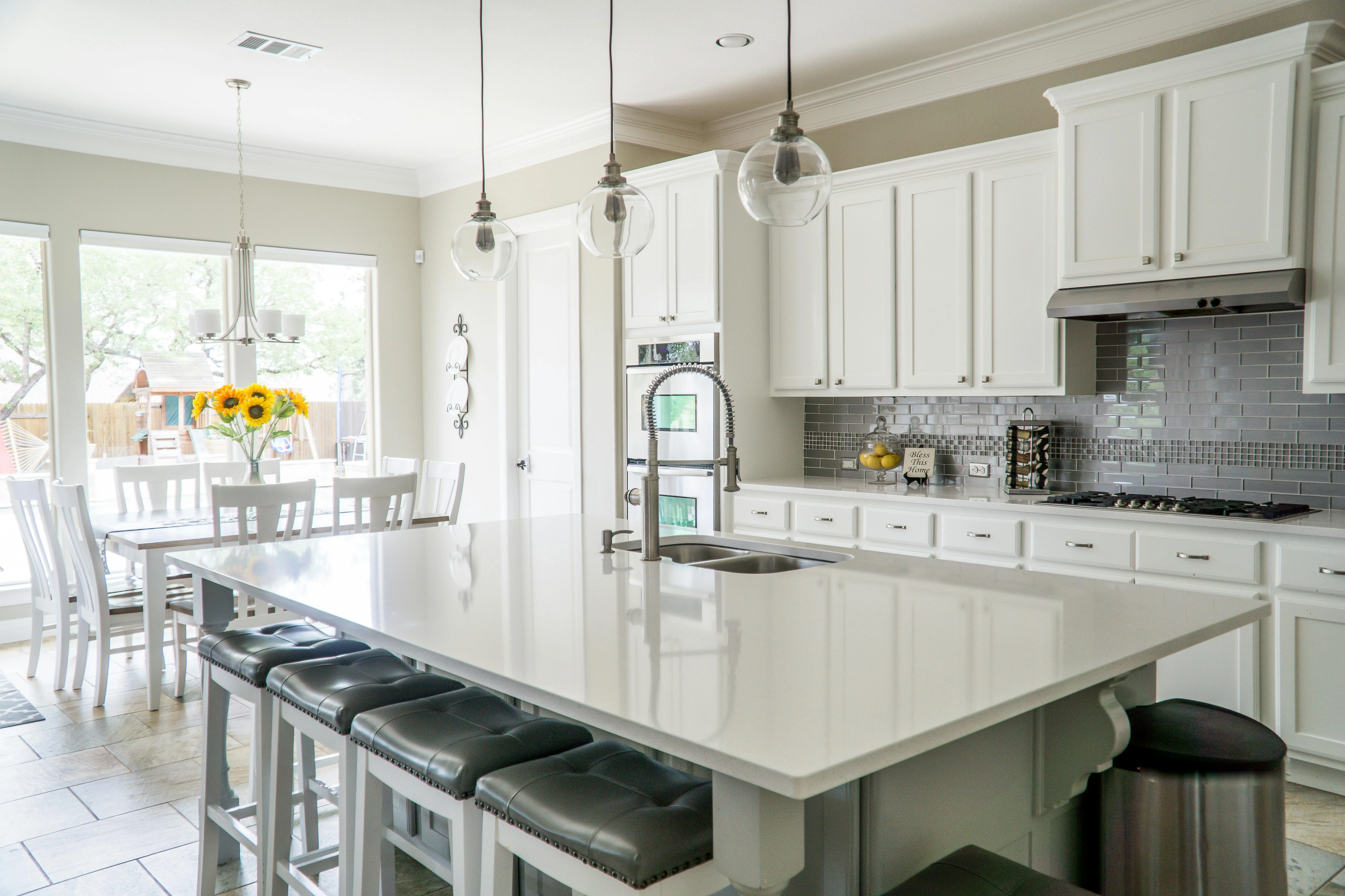 Bright modern kitchen with a large white island, black cushioned stools, white cabinets, stainless steel appliances, gray subway tile backsplash, and a dining table with sunflowers.