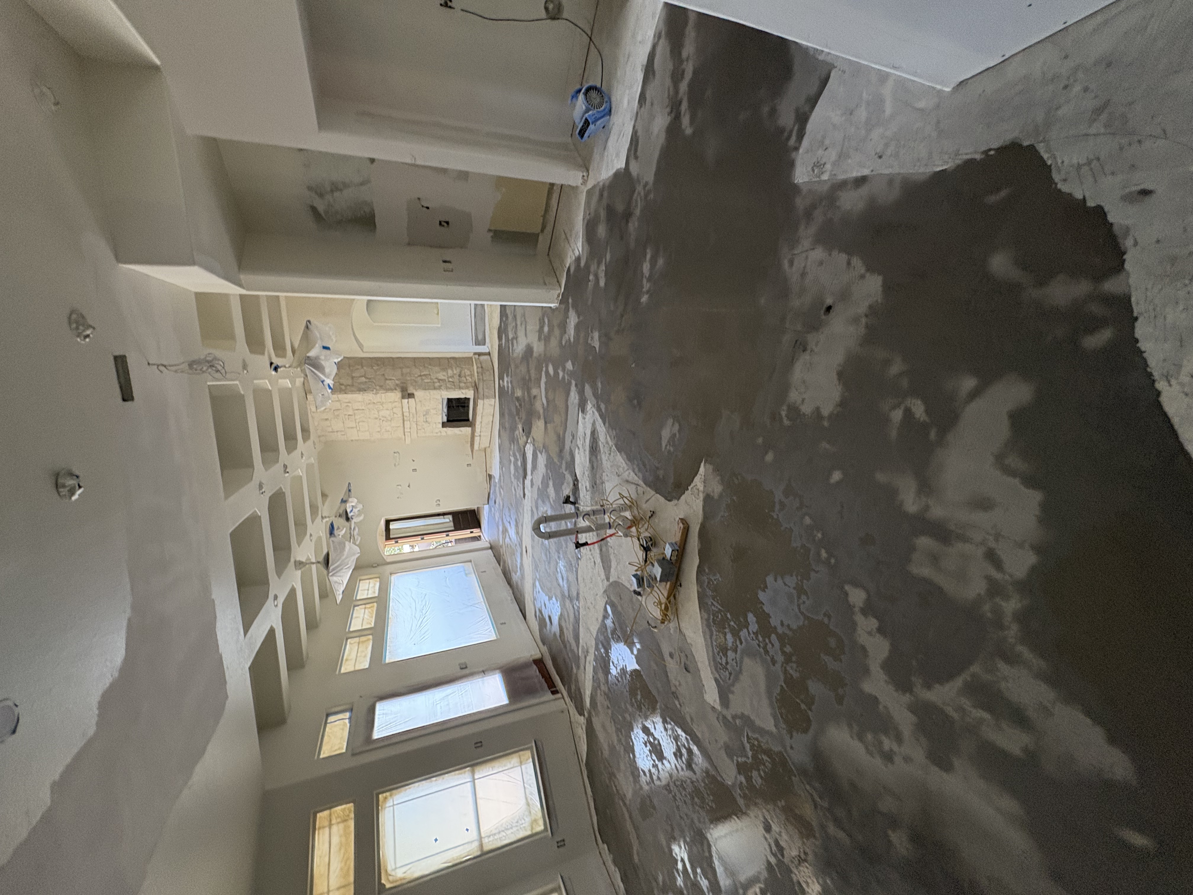 Interior of a large room under renovation with wet concrete floor, taped windows, ceiling fans covered in plastic, and a stone fireplace.