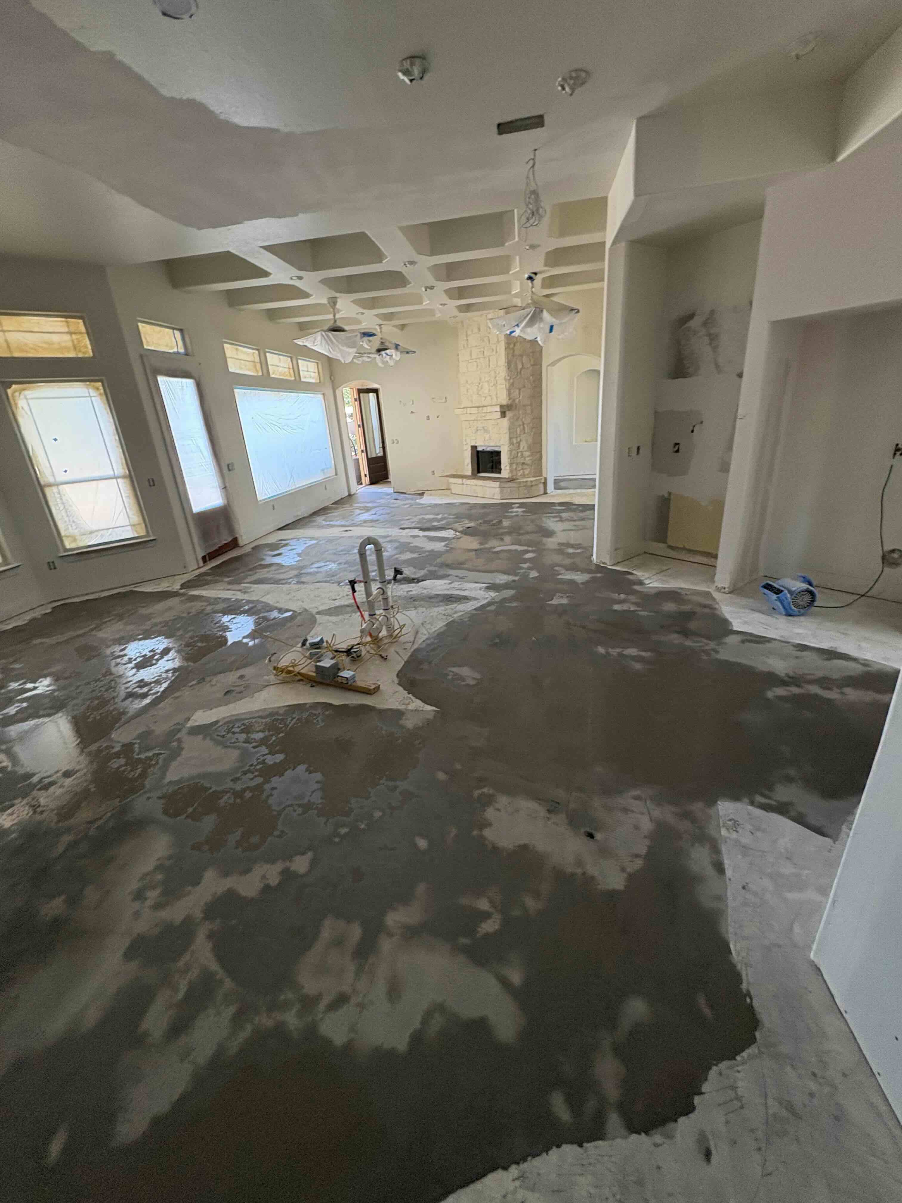 Interior of a large room under renovation with wet concrete floor, taped windows, ceiling fans covered in plastic, and a stone fireplace.