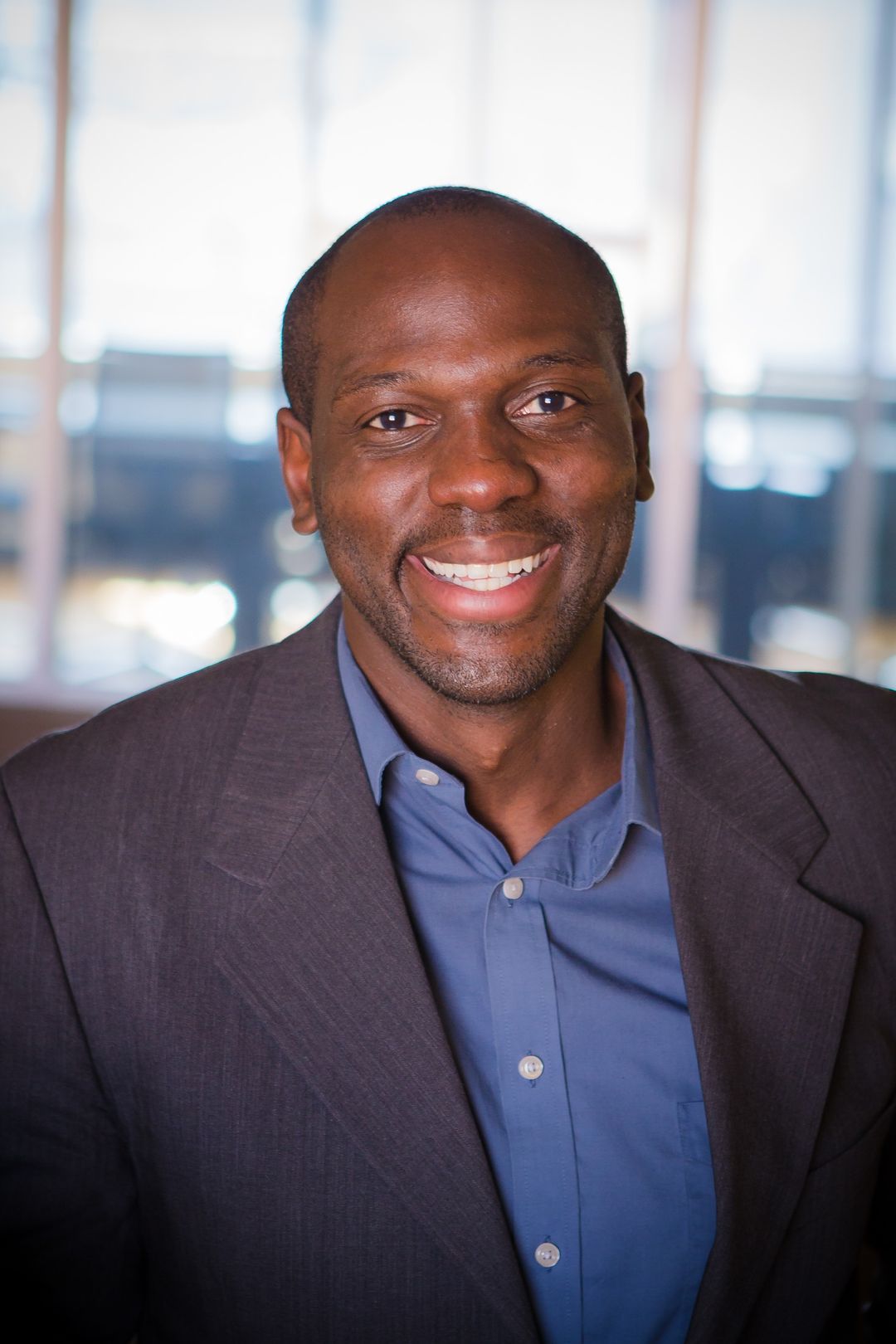 Charcounting CEO smiling and wearing a dark blazer and blue button-up shirt in an office setting with blurred windows in the background.