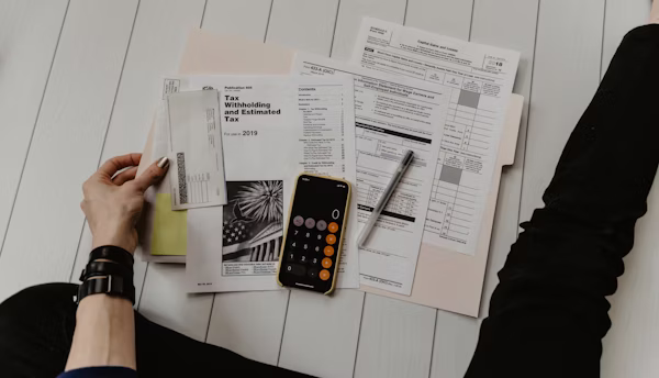 Person sitting on a white floor with tax documents, a calculator app open on a smartphone, and a pen arranged on a folder.