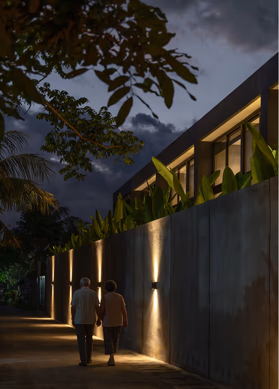 An elderly couple walking hand in hand along a dimly lit street at dusk, beside a modern building with wall-mounted lights and lush greenery above the wall.